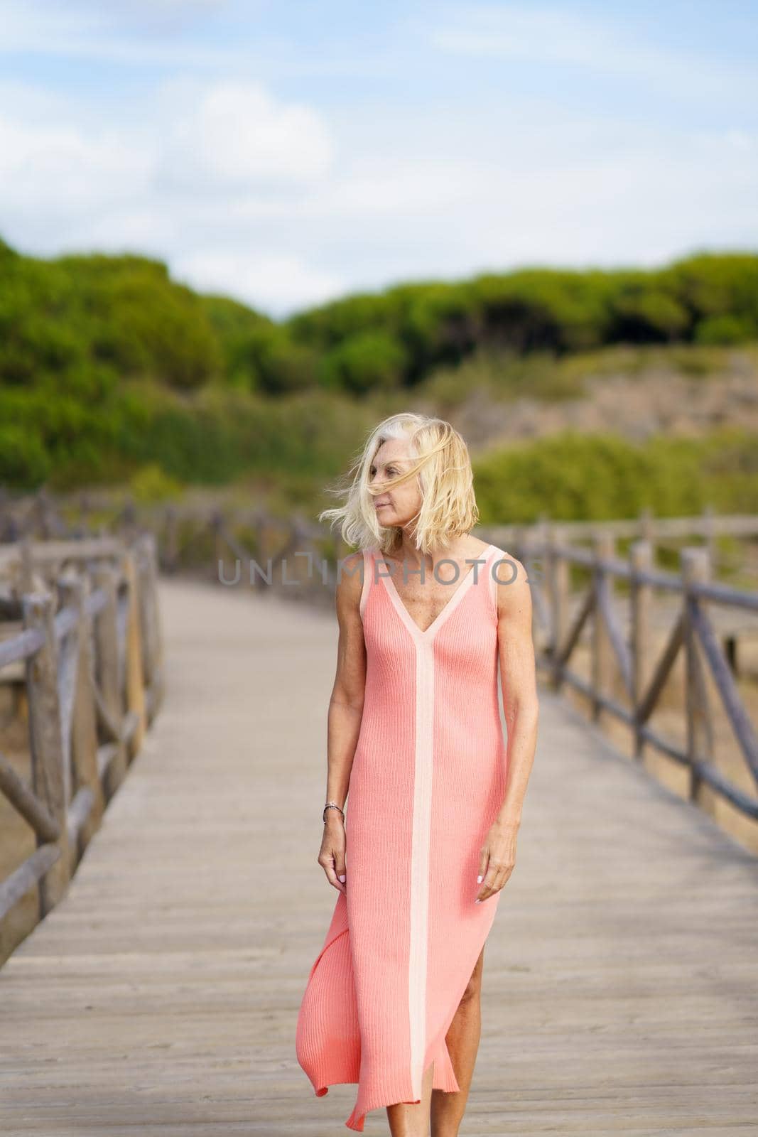 Beautiful mature woman walking along a wooden path near the beach., wearing a nice orange dress. Elderly female enjoying her retirement at a seaside retreat.