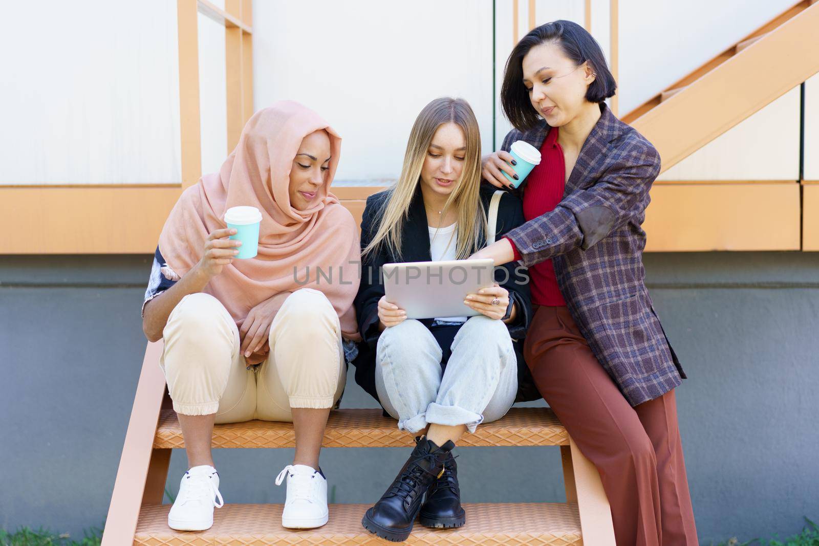 Stylish diverse women friends sharing tablet and drinking takeaway coffee on street by javiindy