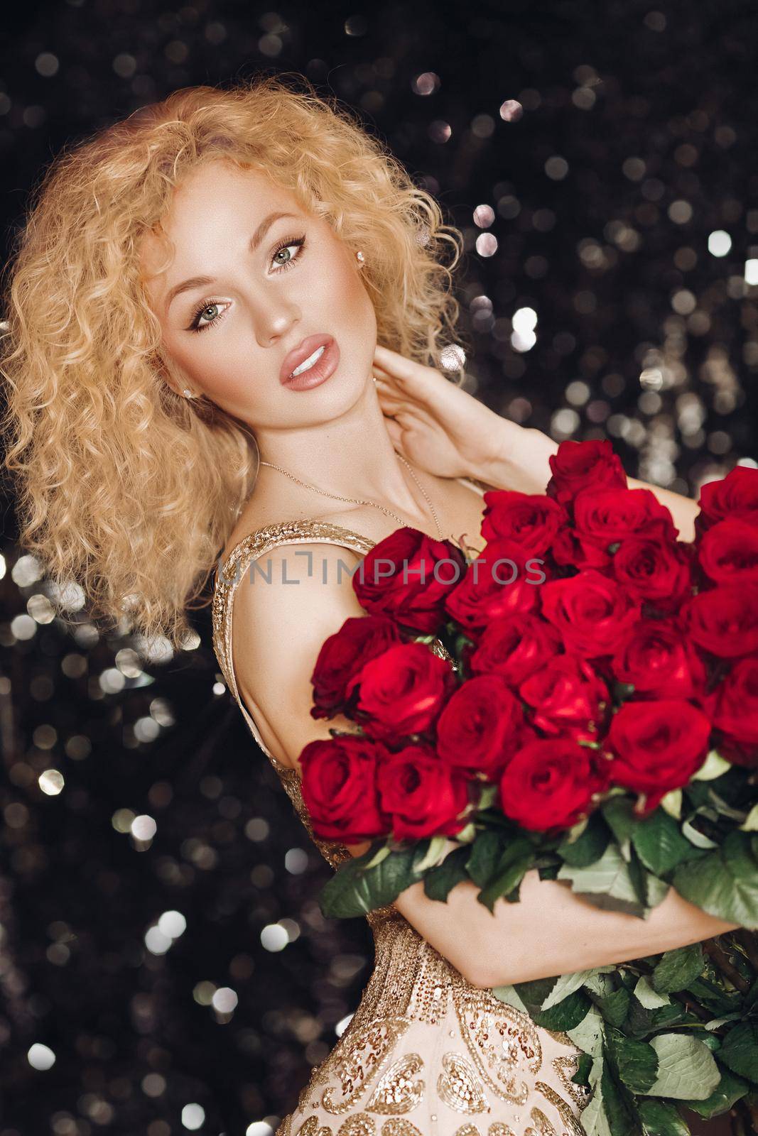 Portrait of gorgeous young blonde woman with curly hair and nude make-up in expensive elegant dress looking at camera holding beautiful red roses. Sparkling background. Bokeh.