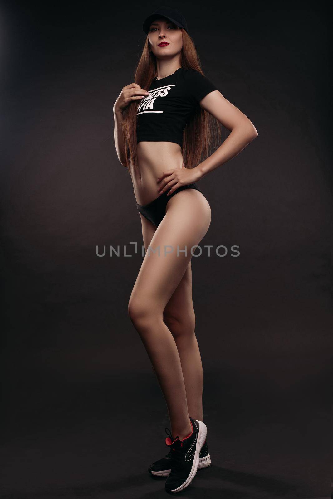 Sexy slim model in top, cap and sneakers posing and looking at camera on black isolated background in studio. Attractive woman doing sport and fitness, caring body shape. Concept of power and fit.