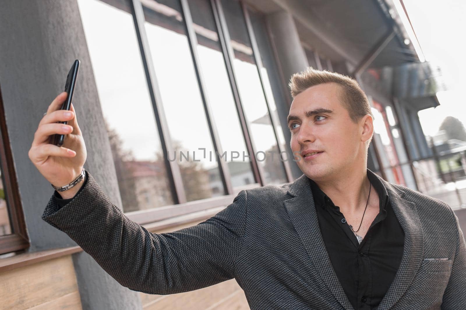 Businessman man of beautiful European appearance, in a jacket and shirt makes a selfie or talks on a video call on a smartphone on the street outdoor.