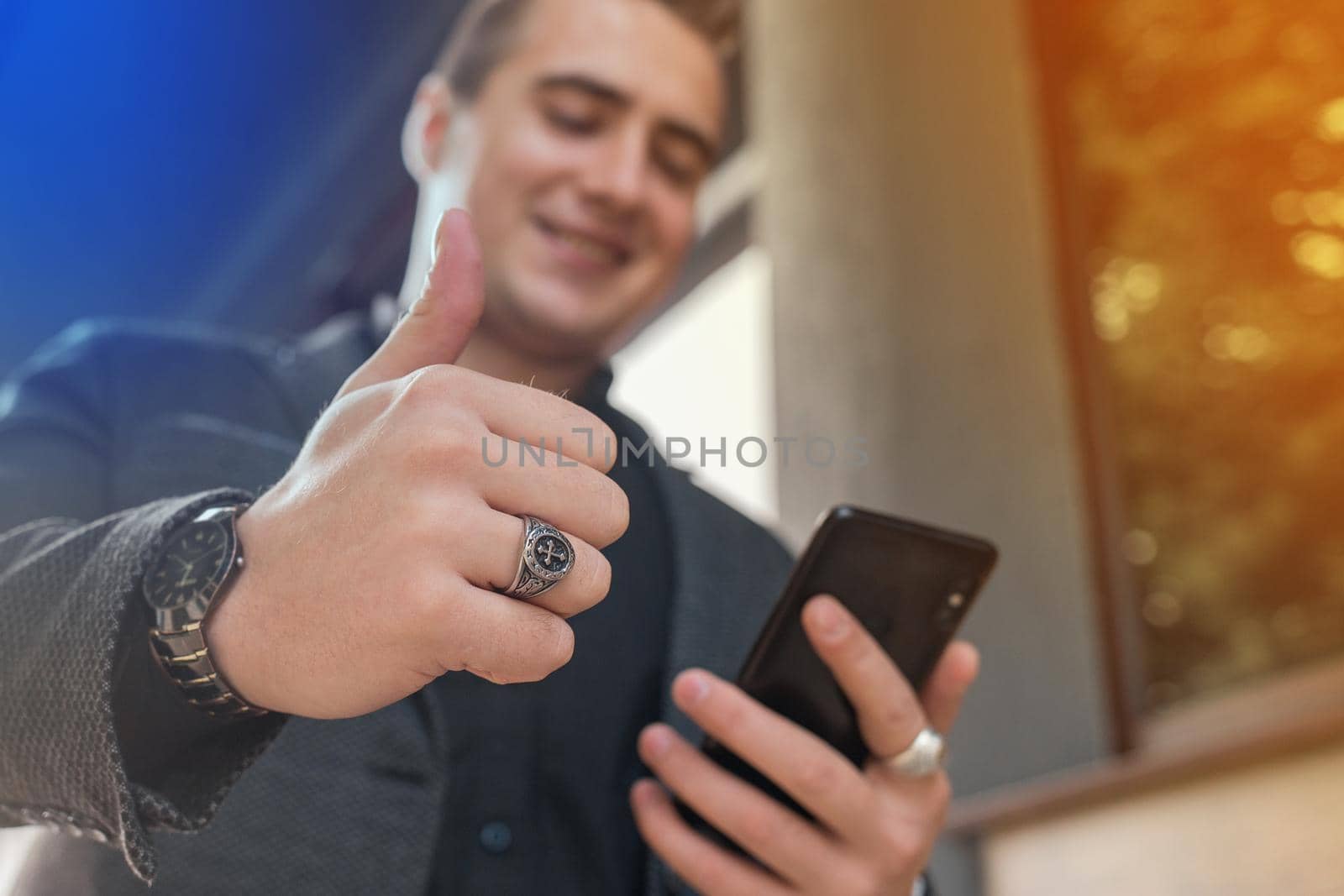 A young positive man in a grey jacket and black shirt holds a mobile phone or smartphone and points the class with his finger up on the street outdoor, close-up by AYDO8