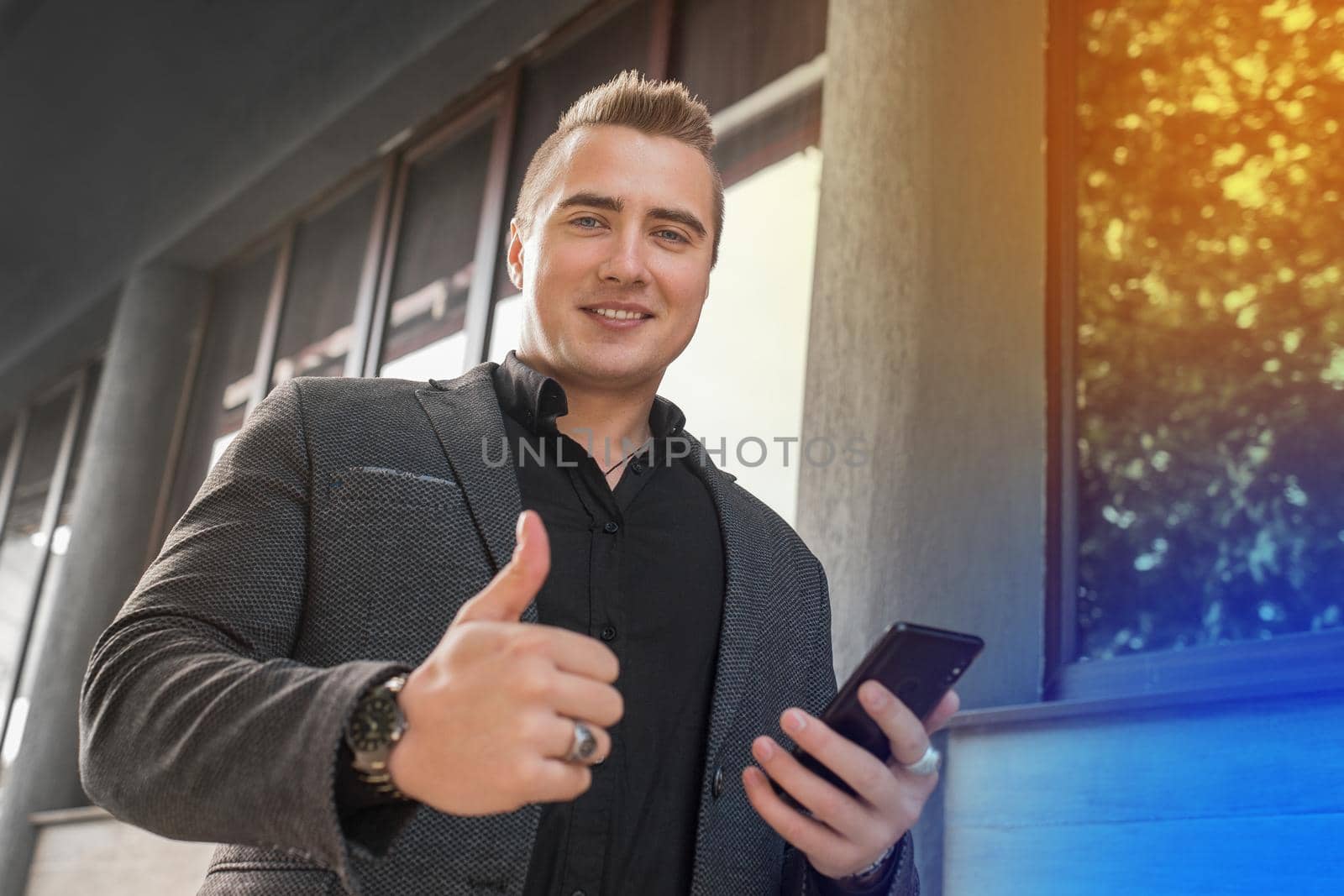 A portrait of a cheerful stylish young guy of Caucasian appearance in a jacket and shirt uses a smartphone and shows the class with a finger up on the outdoor street. close-up.