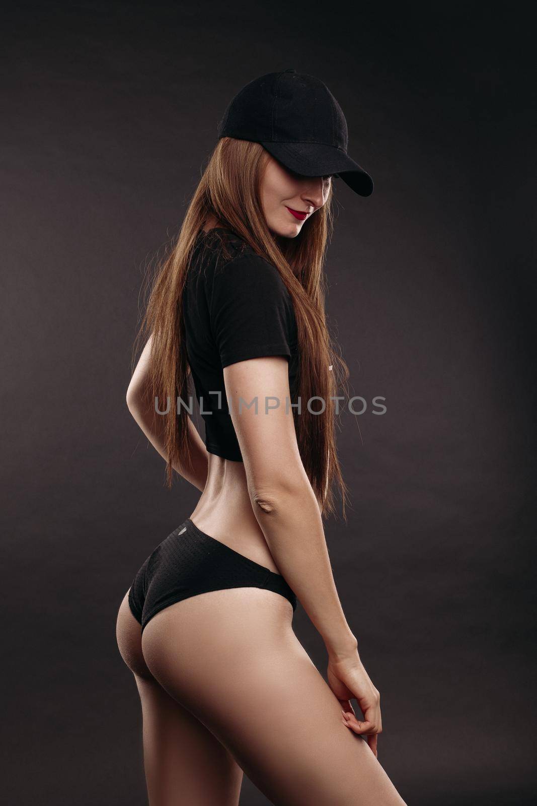 Sexy slim model in top, cap and sneakers posing and looking at camera on black isolated background in studio. Attractive woman doing sport and fitness, caring body shape. Concept of power and fit.