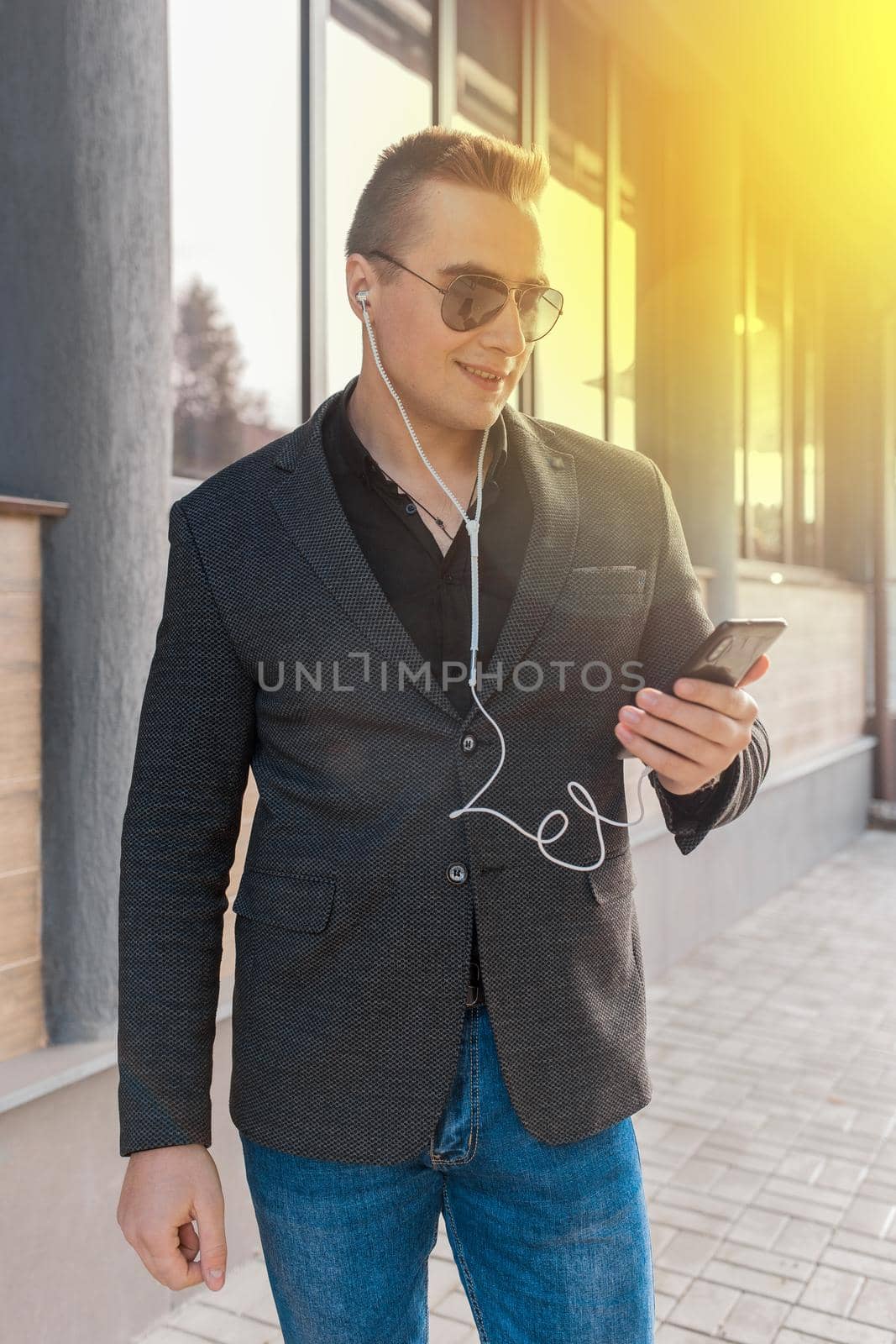 A young attractive businessman of European appearance in a jacket and shirt style, wears sunglasses and listens to music in headphones from a smartphone on the street outdoor.