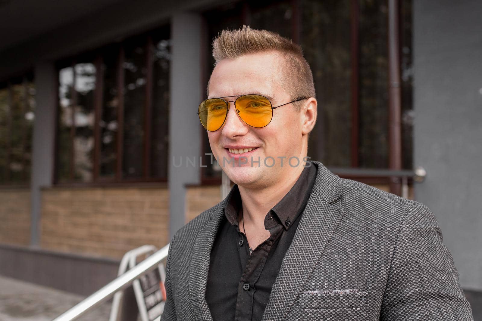 Smiling stylish young guy businessman of European appearance in a gray jacket and black shirt portrait in glasses on the street outdoor.