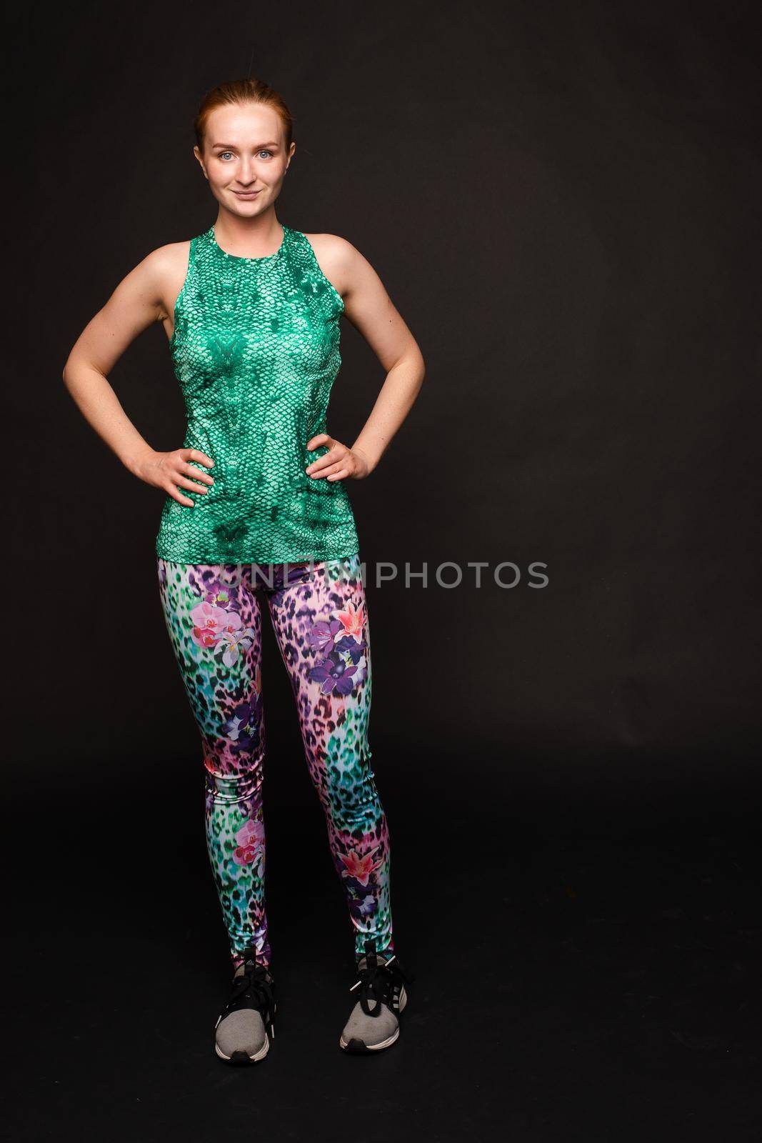 Front view of strong red haired woman in green uniform looking at camera and posing on black isolated background. Muscular girl keeping fit and training. Concept of health and workout.