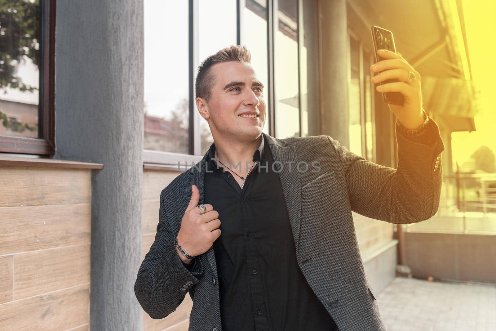 Businessman smiling man portrait of beautiful European appearance, in a jacket and shirt makes a selfie or talks on a video call on a smartphone on the street outdoor by AYDO8