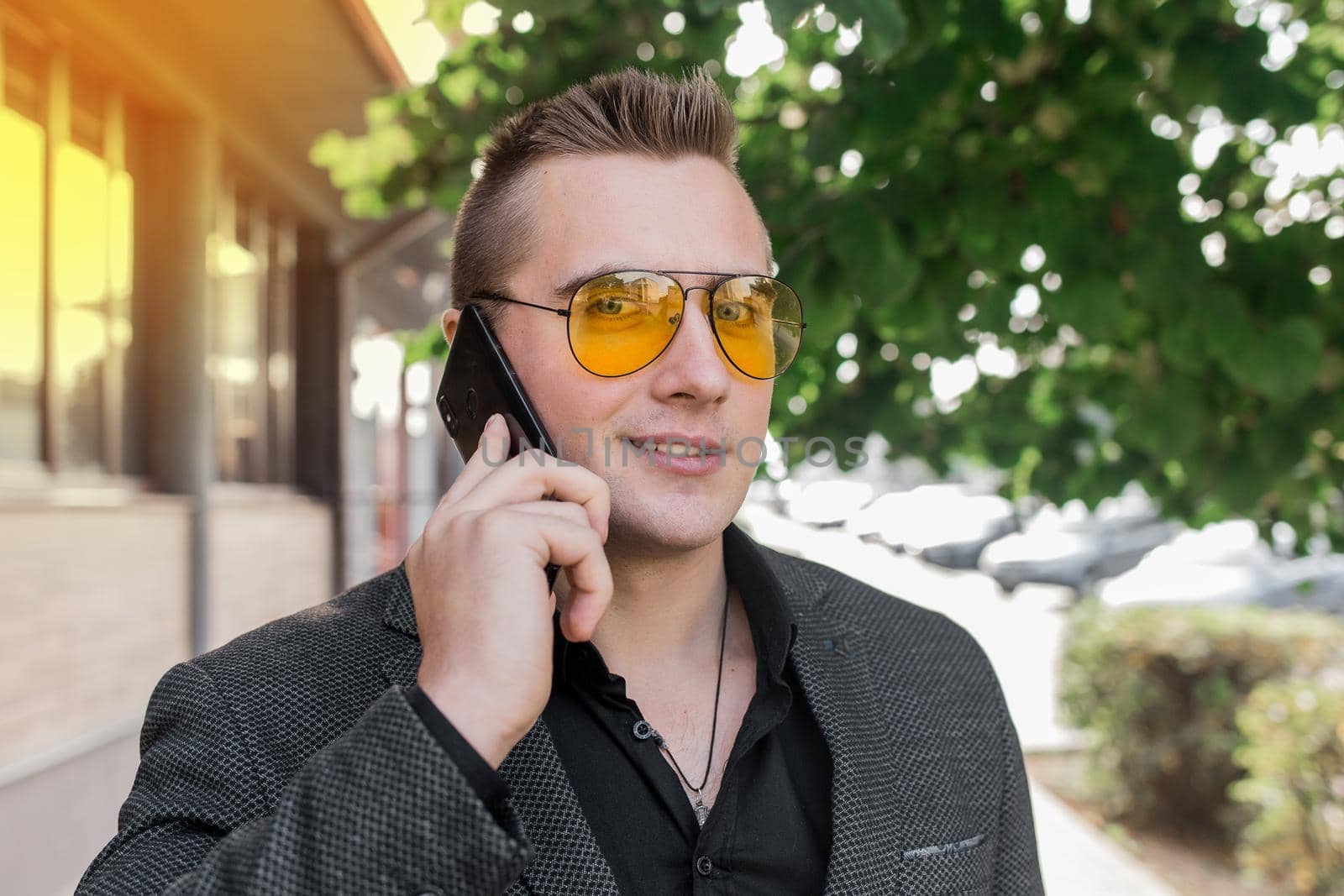 Stylish young positive businessman of attractive European appearance in sunglasses, jacket and shirt, talking on a mobile phone on the outdoor street.