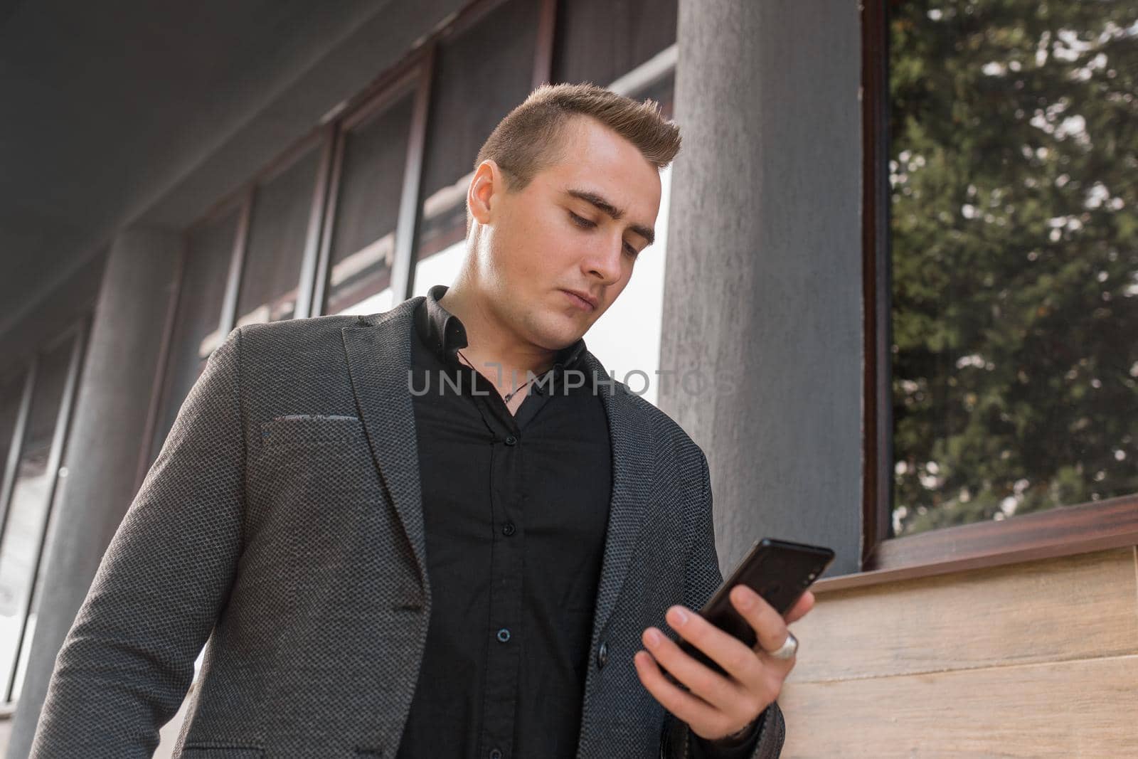 A portrait of a stylish young guy of Caucasian appearance in a jacket and shirt uses a smartphone on the outdoor street. close-up by AYDO8