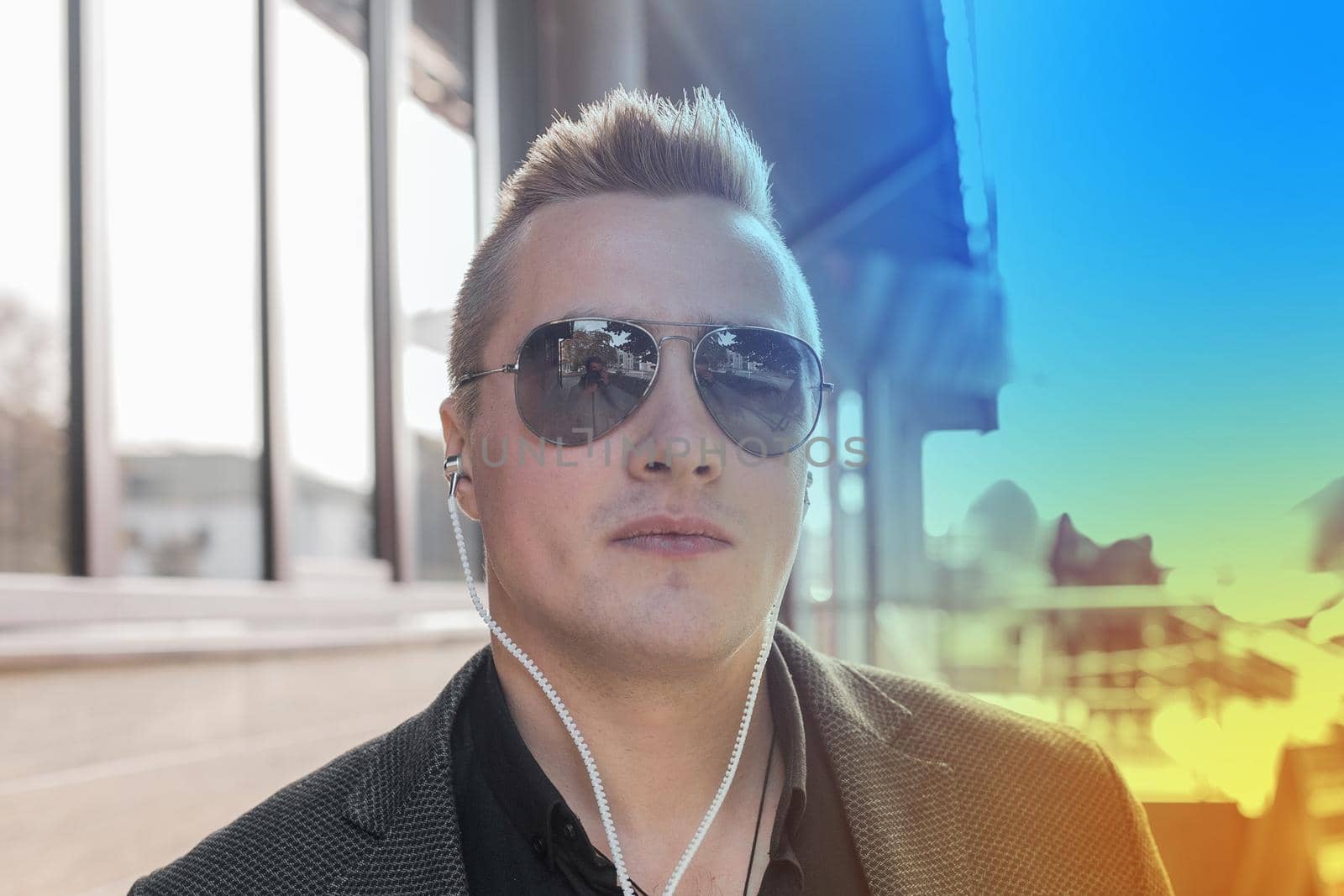 Portrait of a serious young guy of European appearance, an attractive businessman in sunglasses, listening to music in headphones on the street outdoor by AYDO8