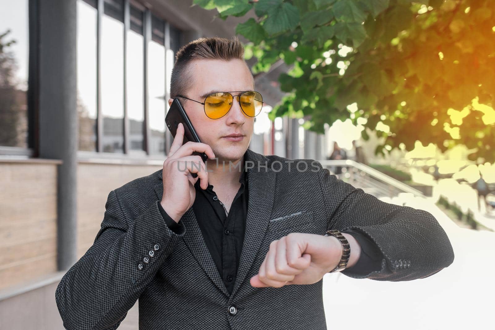 A young serious guy, businessman of European appearance portrait in sunglasses talks on a mobile phone or smartphone and looks at the time on a hand watch on the street outdoor.