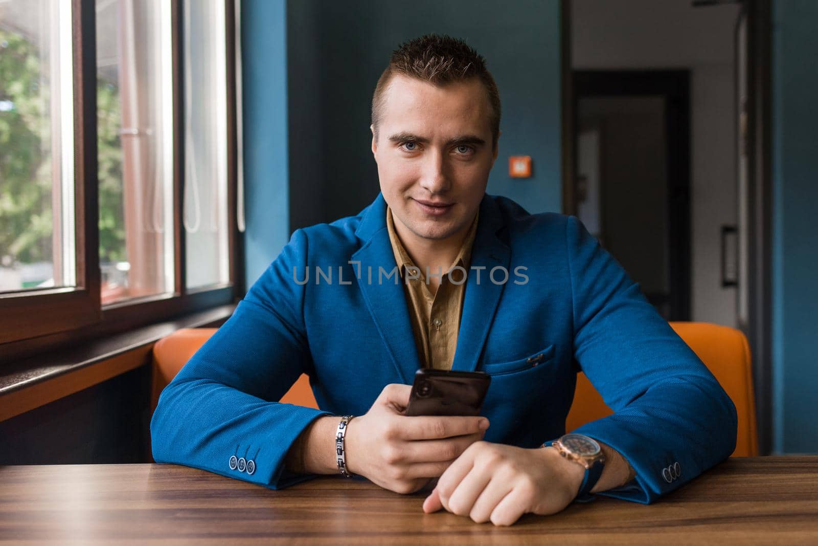 A satisfied man of European appearance portrait of an attractive businessman, in a blue jacket and shirt uses a smartphone sitting at a table in a cafe.