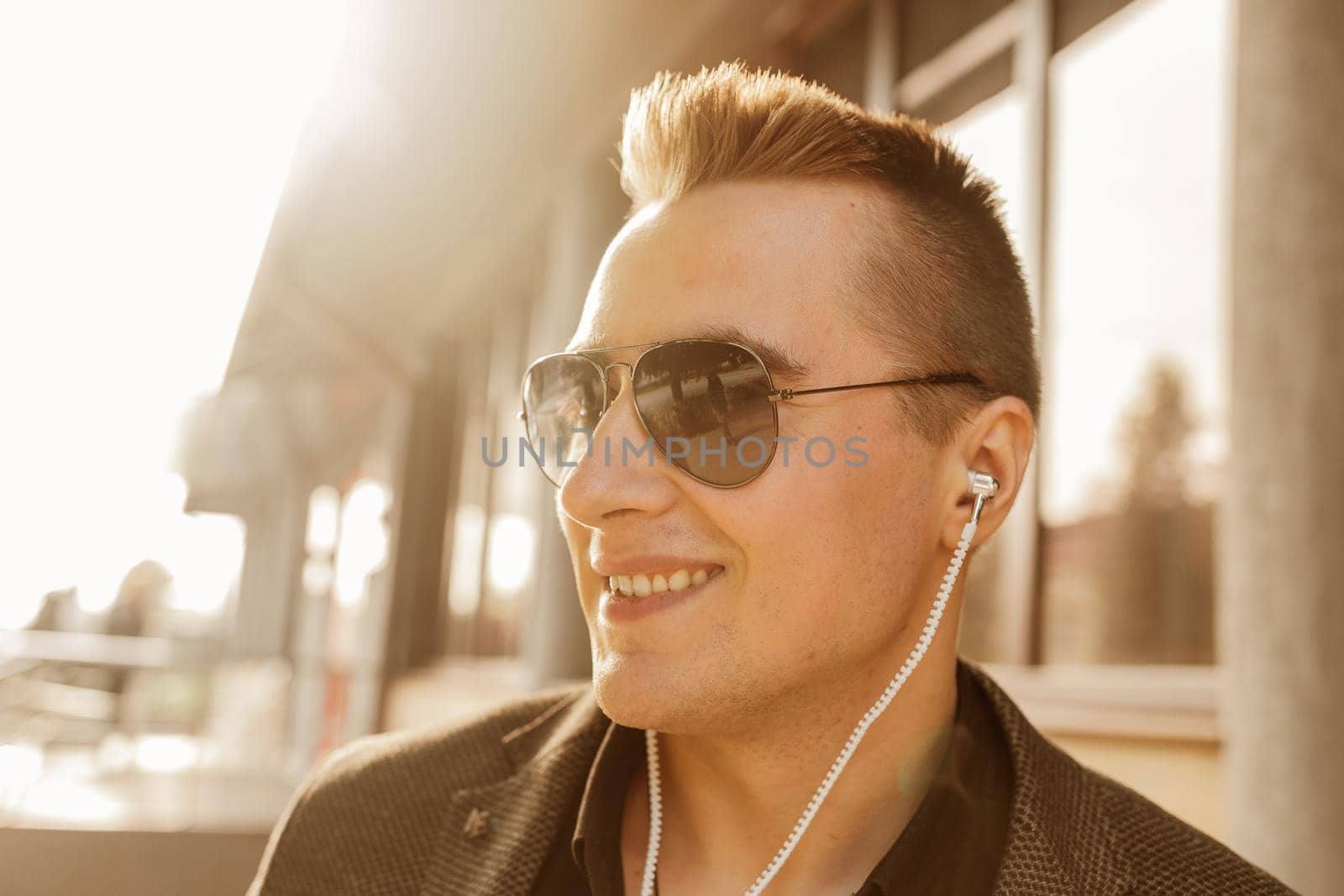 Portrait of a positive smiling young businessman of European appearance in sunglasses and headphones listening to music on the street outdoor by AYDO8