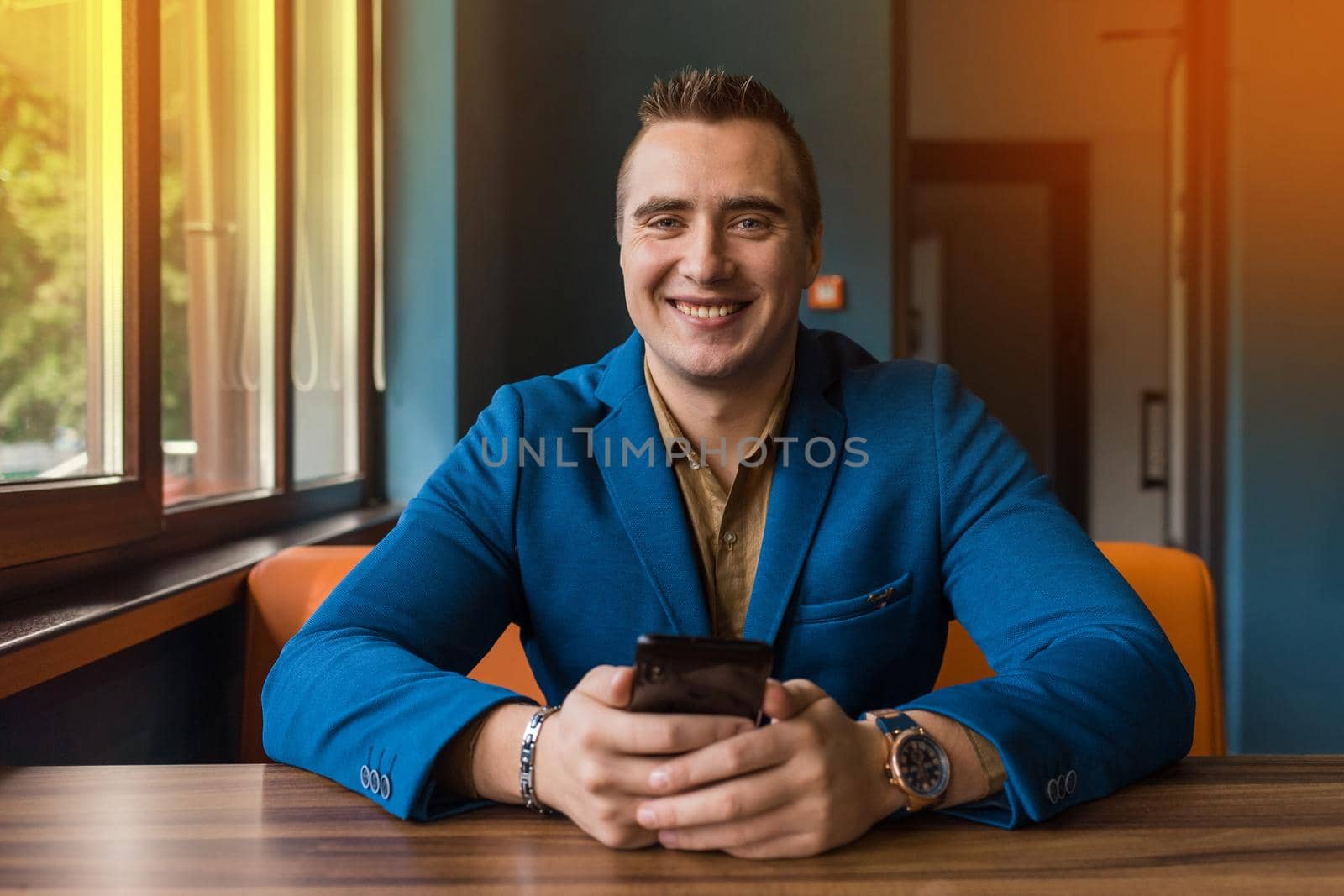 Smiling stylish young, businessman of European appearance portrait of a man in a jacket and shirt sits at a table and uses a mobile phone in a cafe.