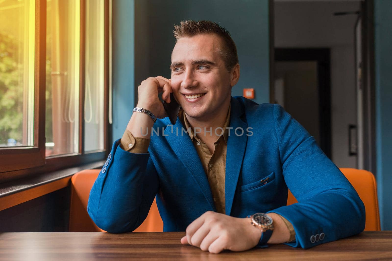 An adult stylish, smiling positive businessman of European appearance portrait sits in a blue jacket at a table in a cafe and talks on a smartphone looking out the window.