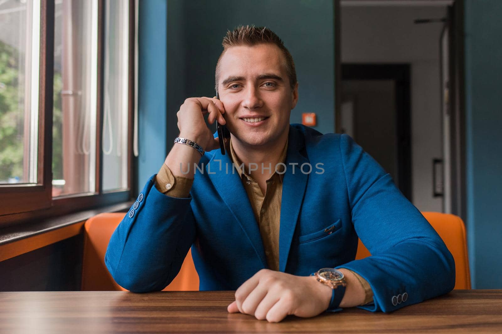 An adult stylish, smiling positive businessman of European appearance portrait sits in a jacket at a table in a cafe and talks on a smartphone or mobile phone.