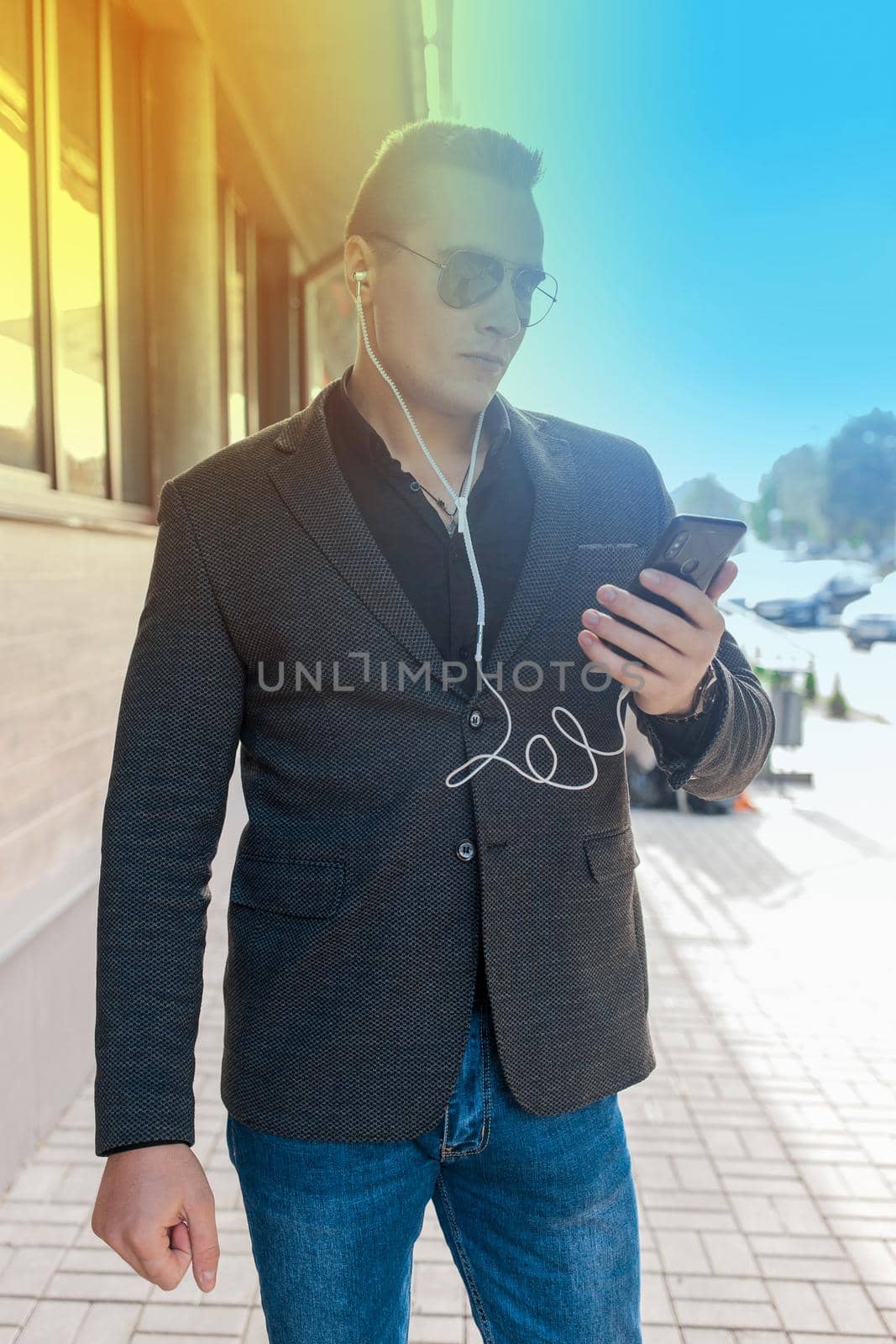 Portrait of a serious man of European appearance in a jacket, shirt and jeans, in sunglasses, with headphones and a smartphone in his hands on the street outdoor by AYDO8