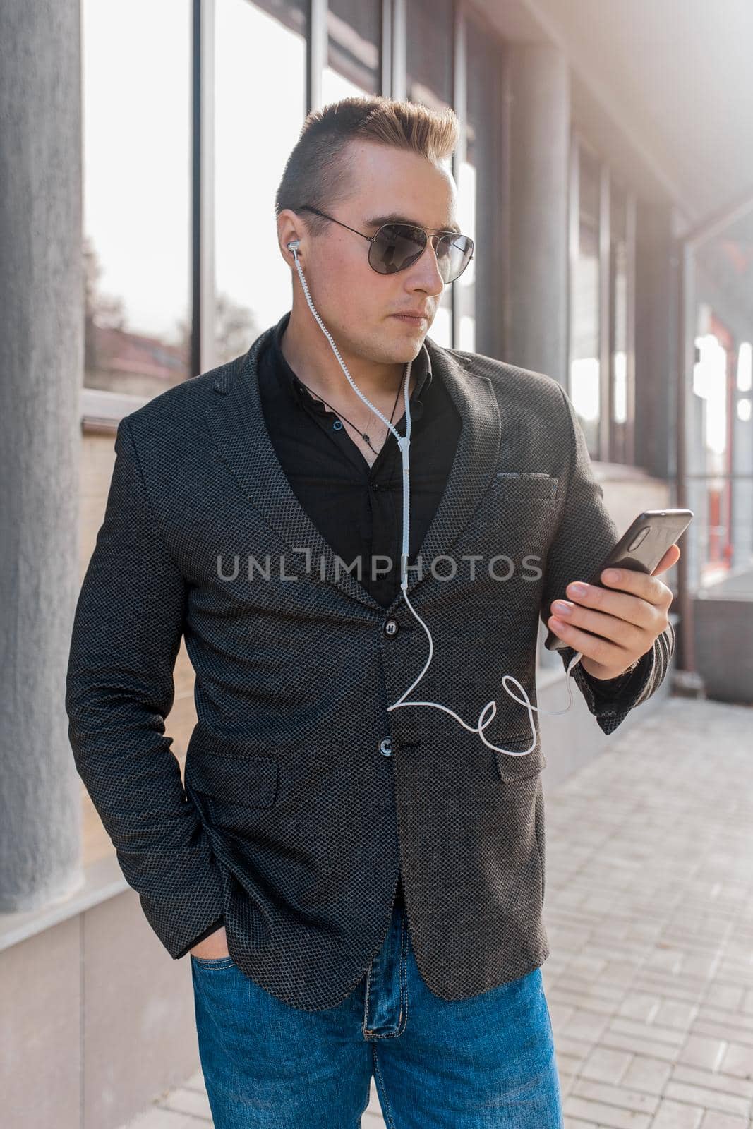 Portrait of a young business man in sunglasses, jacket and shirt with jeans and headphones listening to music from a mobile phone in the street outdoors.