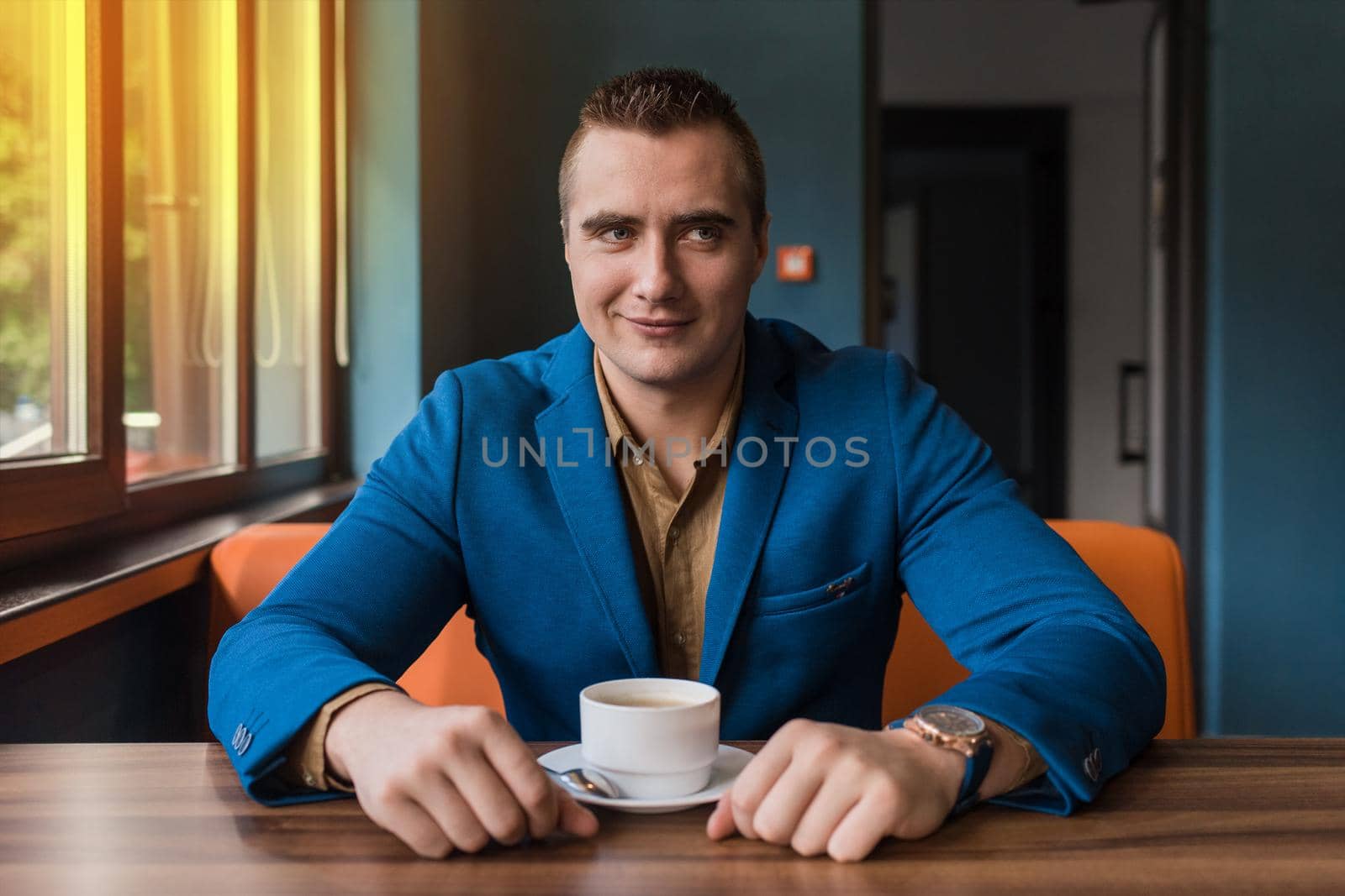 A adult, businessman portrait of Caucasian appearance in a blue jacket and brown shirt sits at a table in a cafe with a cup of coffee on a break.