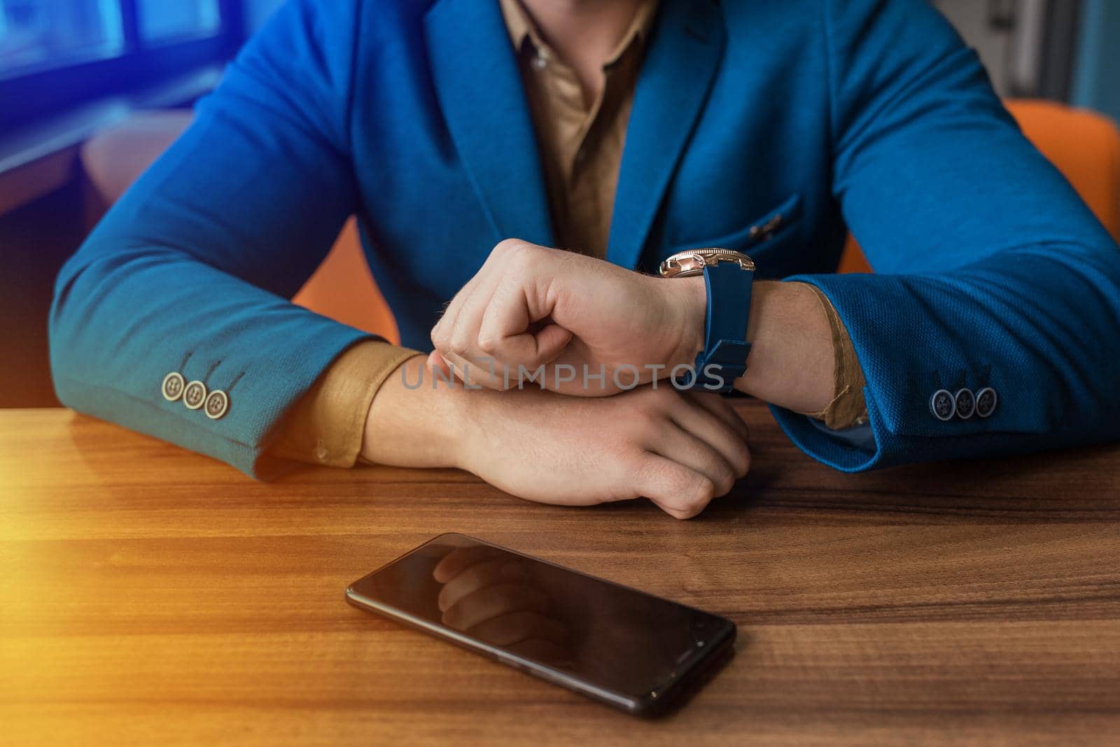 A businessman in a blue jacket looks at the time on a hand watch, opposite lies on a wooden table of a mobile phone or smartphone.