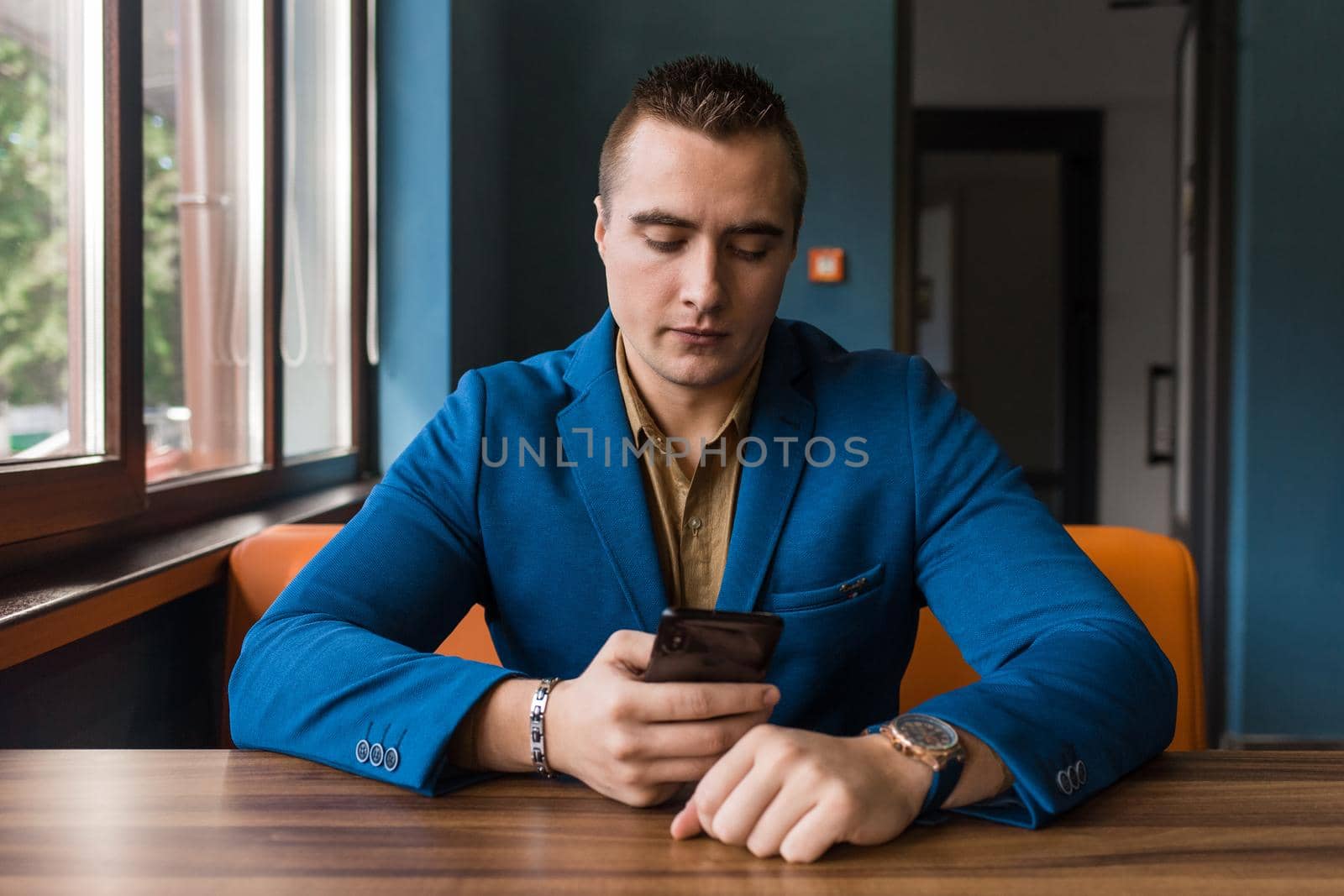 A young businessman of stylish European appearance in a blue suit and brown shirt sits at a table in a cafe in a mobile phone.