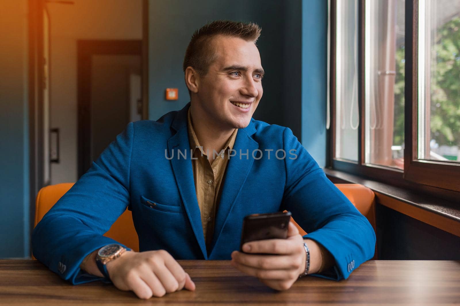 A smiling stylish, young businessman of Caucasian appearance portrait in a jacket and shirt sits at a table in a mobile phone and looks out the window.