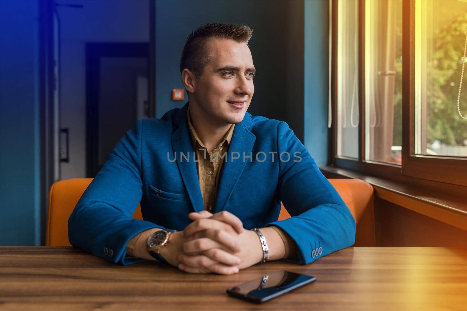 A smiling stylish, young businessman of Caucasian appearance portrait in a jacket and shirt sits at a table in a mobile phone or smartphone and looks out the window.