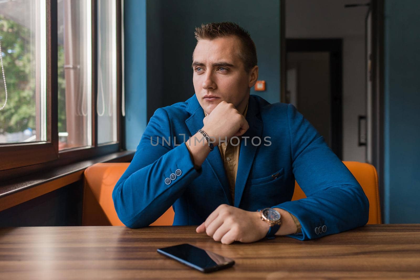 A brooding, stylish businessman portrait of European appearance in a jacket and shirt sits at a table in a cafe for a cup of coffee on a lunch break and looks out the window.