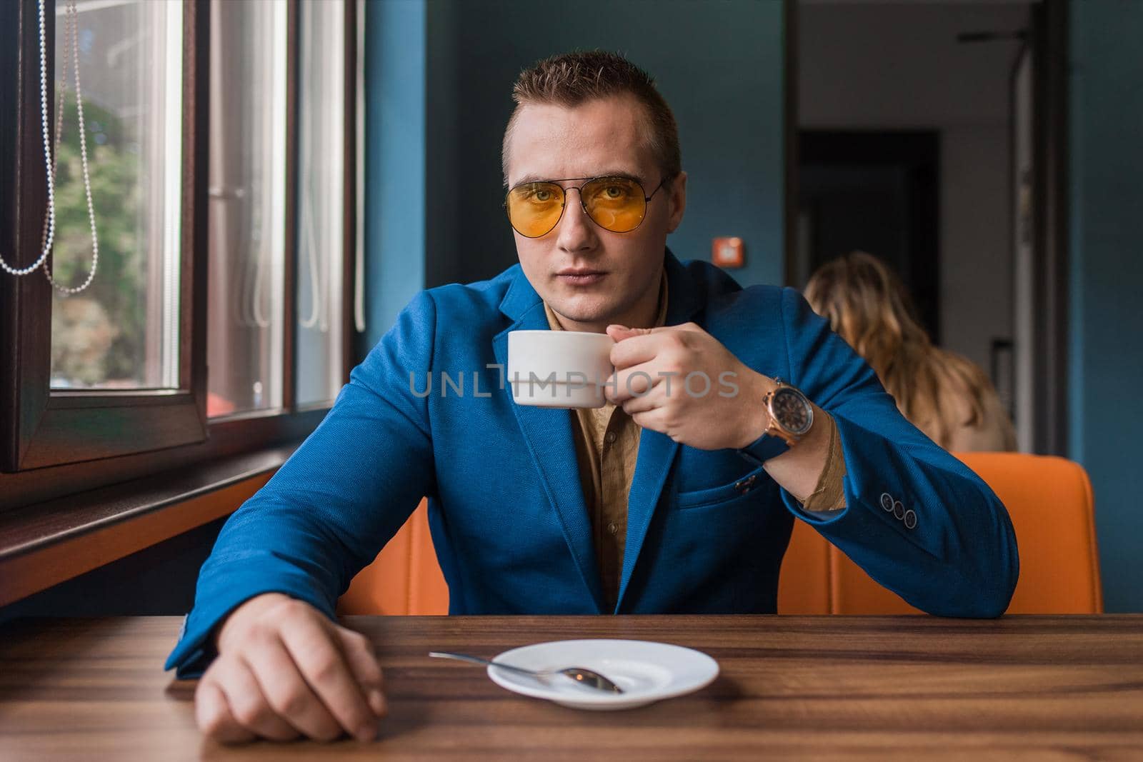 A businessman a stylish portrait of Caucasian appearance in sunglasses, jacket and shirt, sits at a table on a coffee break in a cafe background.