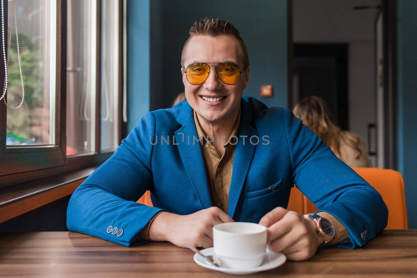 A positive smiling businessman a stylish portrait of Caucasian appearance in sunglasses, blue jacket and brown shirt, sits at a table on a coffee break and looks at camera in a cafe background.