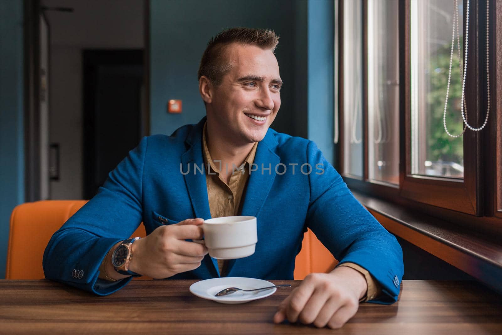 A stylish, positive businessman of Caucasian appearance, a man in a jacket and shirt sits at a table in a cafe on a coffee break and looks away or out the window.