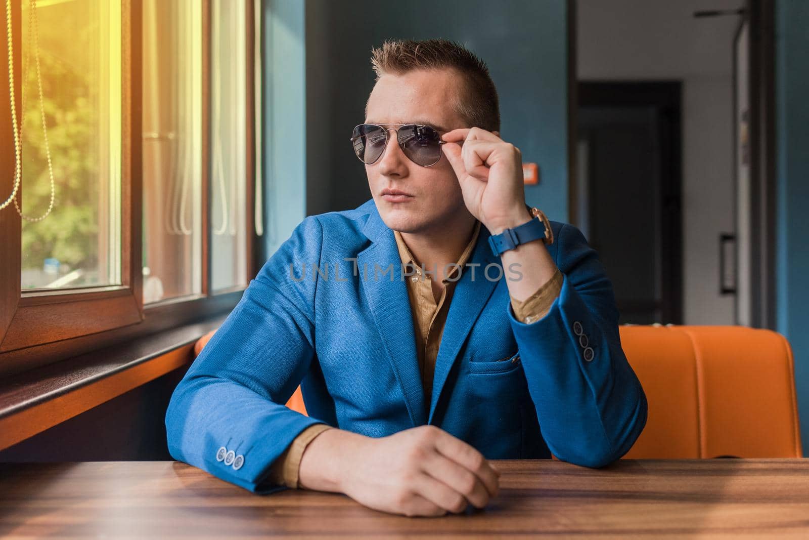 Business young businessman of Caucasian appearance stylish portrait adjusts sunglasses with his hands, looking out the window and sitting at table.