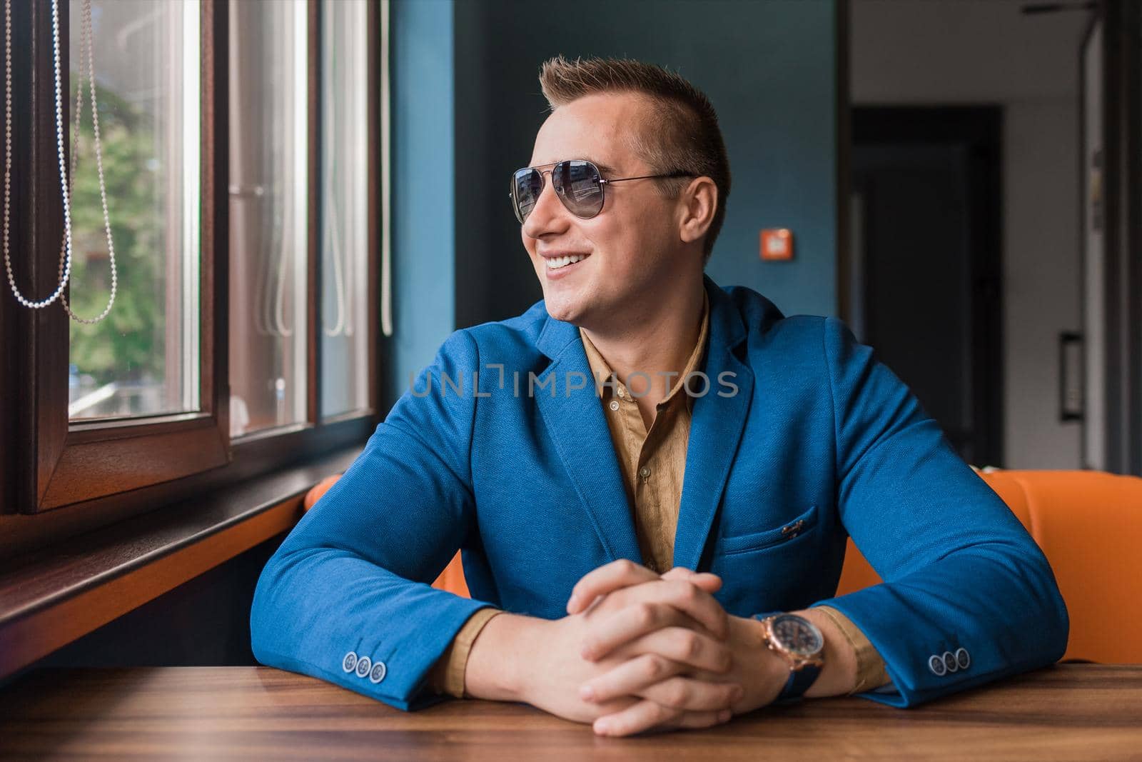 A young positive, smiling businessman of European appearance stylish portrait with sunglasses sits at a table in a cafe idly by at look at the window.