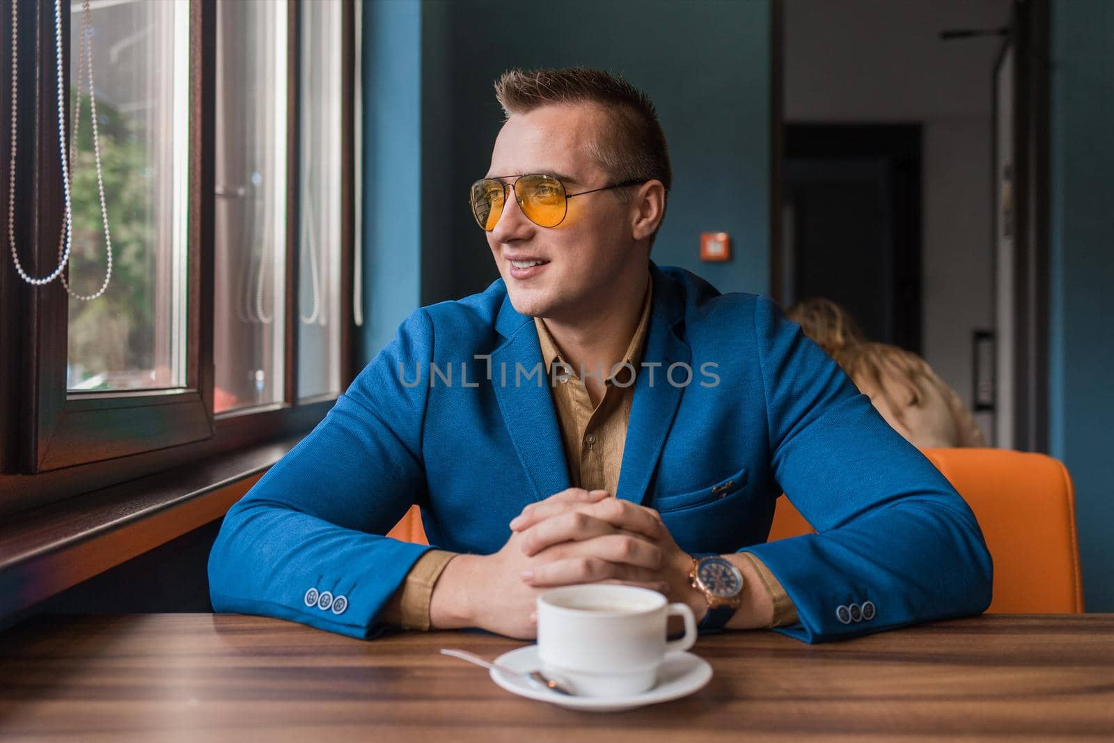 A positive smiling businessman a stylish portrait of Caucasian appearance in sunglasses, sits idly by at a table on a coffee break in a cafe background and looks out the window.