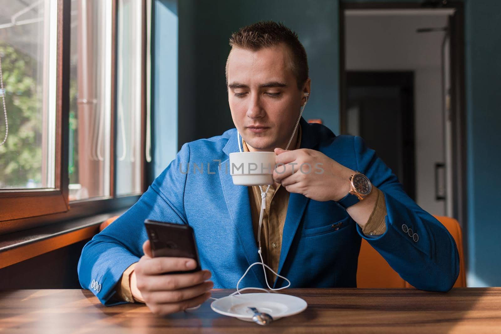 A young handsome guy businessman in blue jacket, of European appearance portrait, uses a smartphone or mobile phone sitting in a cafe at a table on a coffee break.