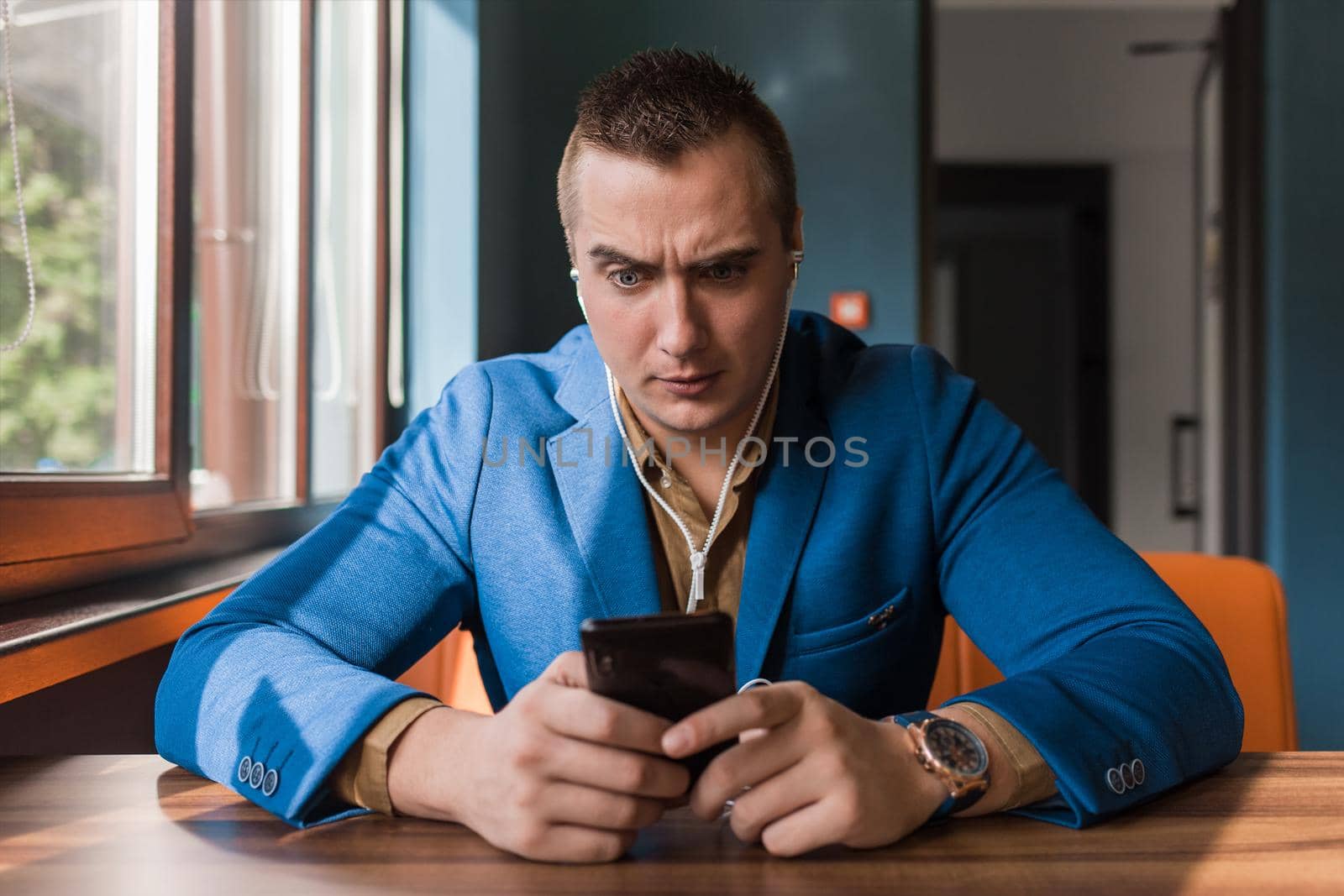 Businessman, a stylish, surprised handsome guy of Caucasian appearance portrait in a blue jacket spends time in a smartphone or mobile phone sitting at a table in the cafe.