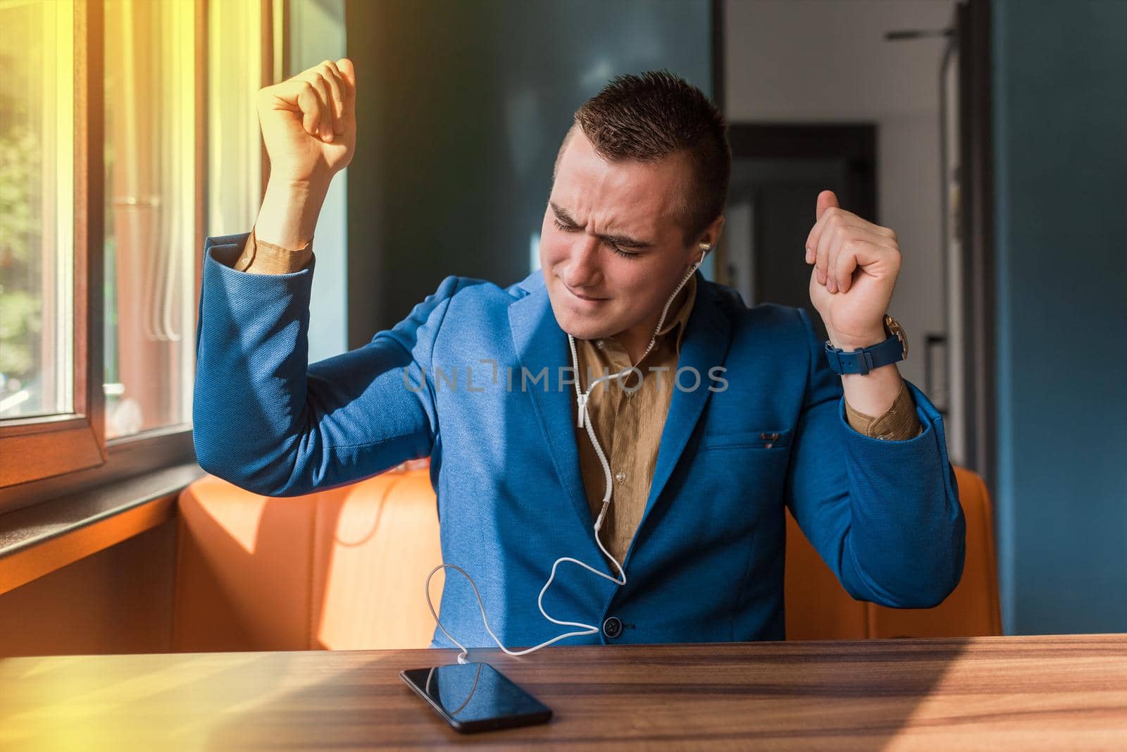 A handsome, happy, funny dancing businessman of European appearance stylish portrait, holds a phone in his hand and listens to music in headphones sitting at a table in a cafe.