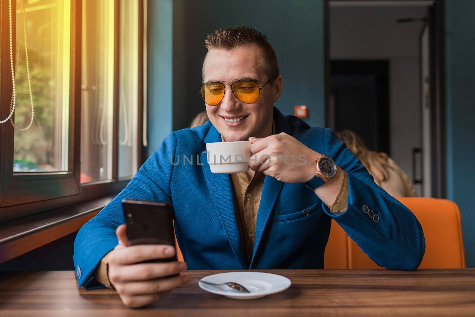 A young smiling positive handsome guy businessman of European appearance portrait, uses a smartphone or mobile phone sitting in a cafe at a table on a coffee break.