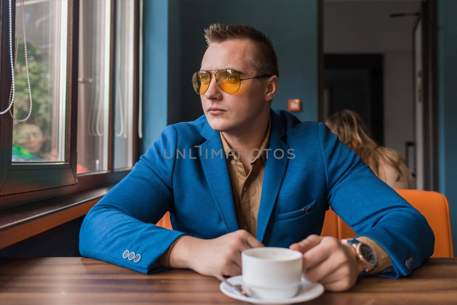 A brooding businessman of young Caucasian appearance, stylish in sunglasses and a suit sits at a table in a cafe over a cup of coffee on a coffee break and looks out the window.