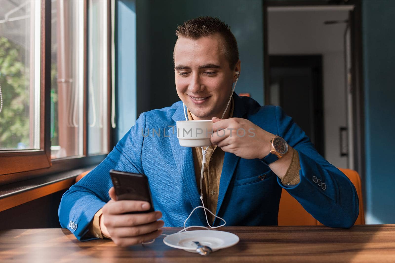A young handsome positive human businessman in blue jacket, of European appearance portrait, uses a smartphone or mobile phone sitting in a cafe at a table on a coffee break.