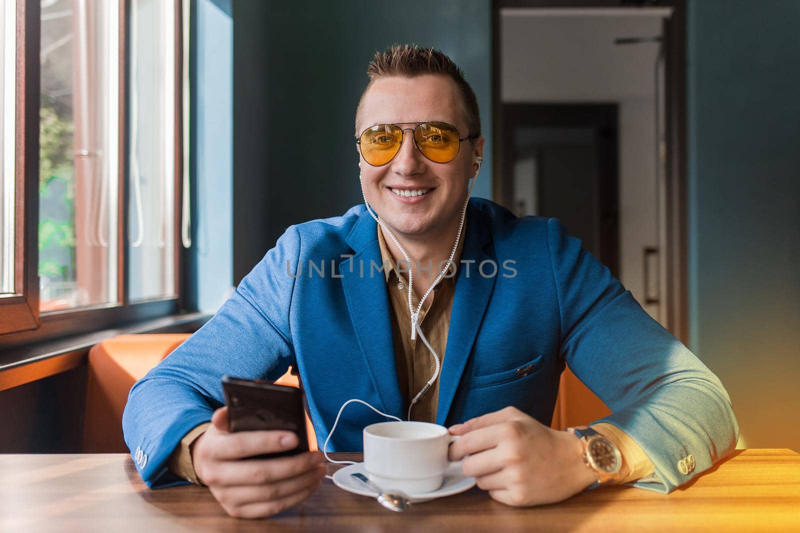 A businessman a stylish portrait of Caucasian appearance in sunglasses, blue suit, sits at a table on a coffee break in smartphone and headphones, listen to music on cellphone a cafe background.
