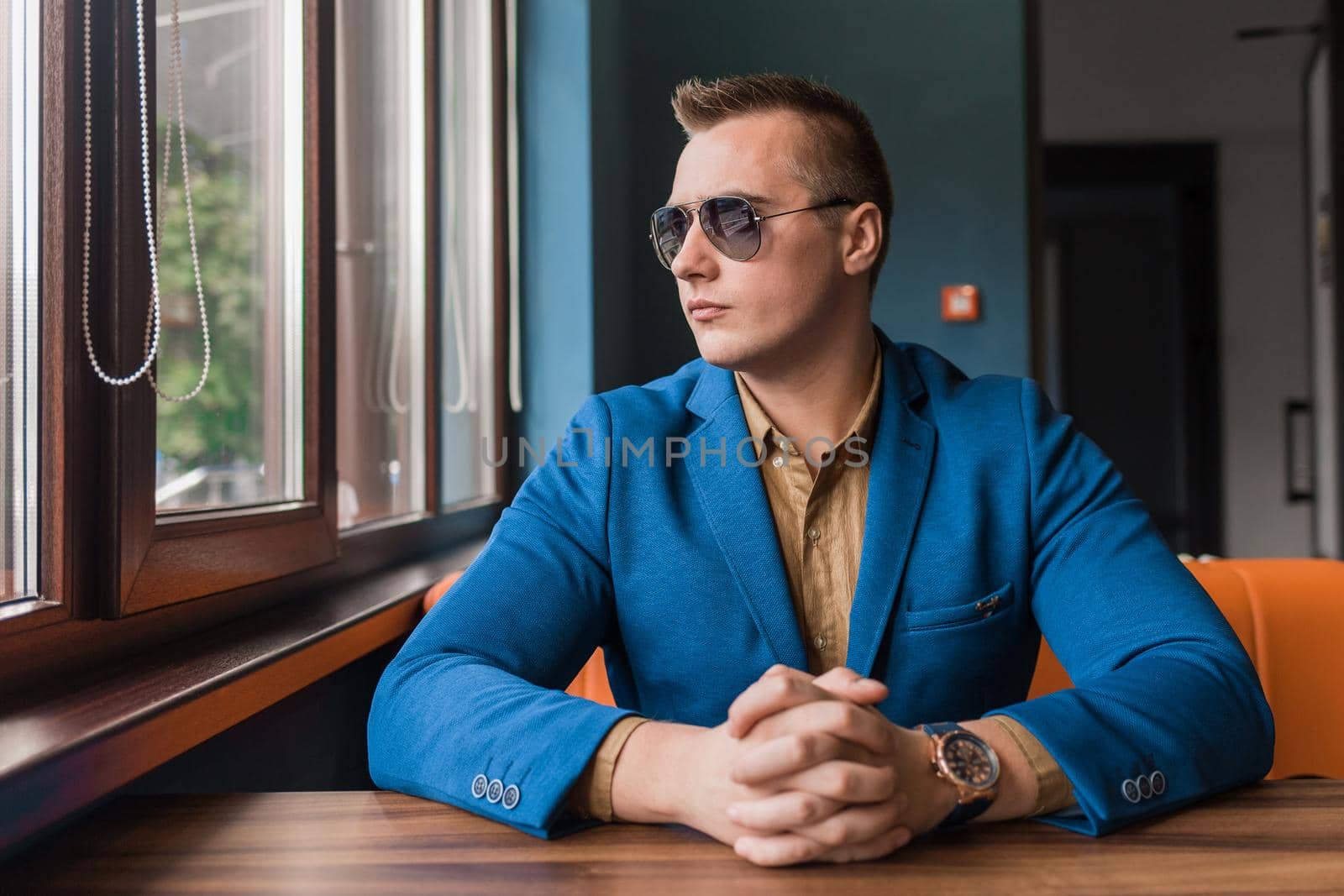 A young businessman of European appearance stylish portrait with sunglasses sits at a table in a cafe idly by at look at the window.