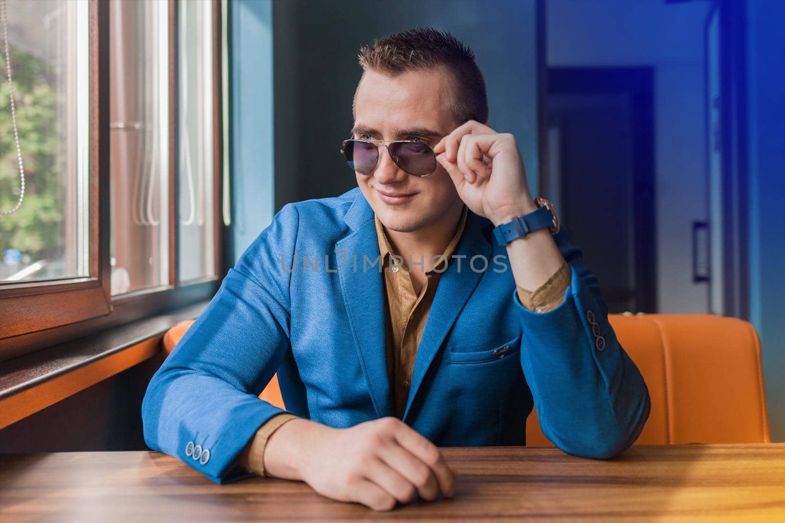 Business young businessman of Caucasian appearance stylish portrait adjusts sunglasses with his hands, looking out the window and sitting at table.