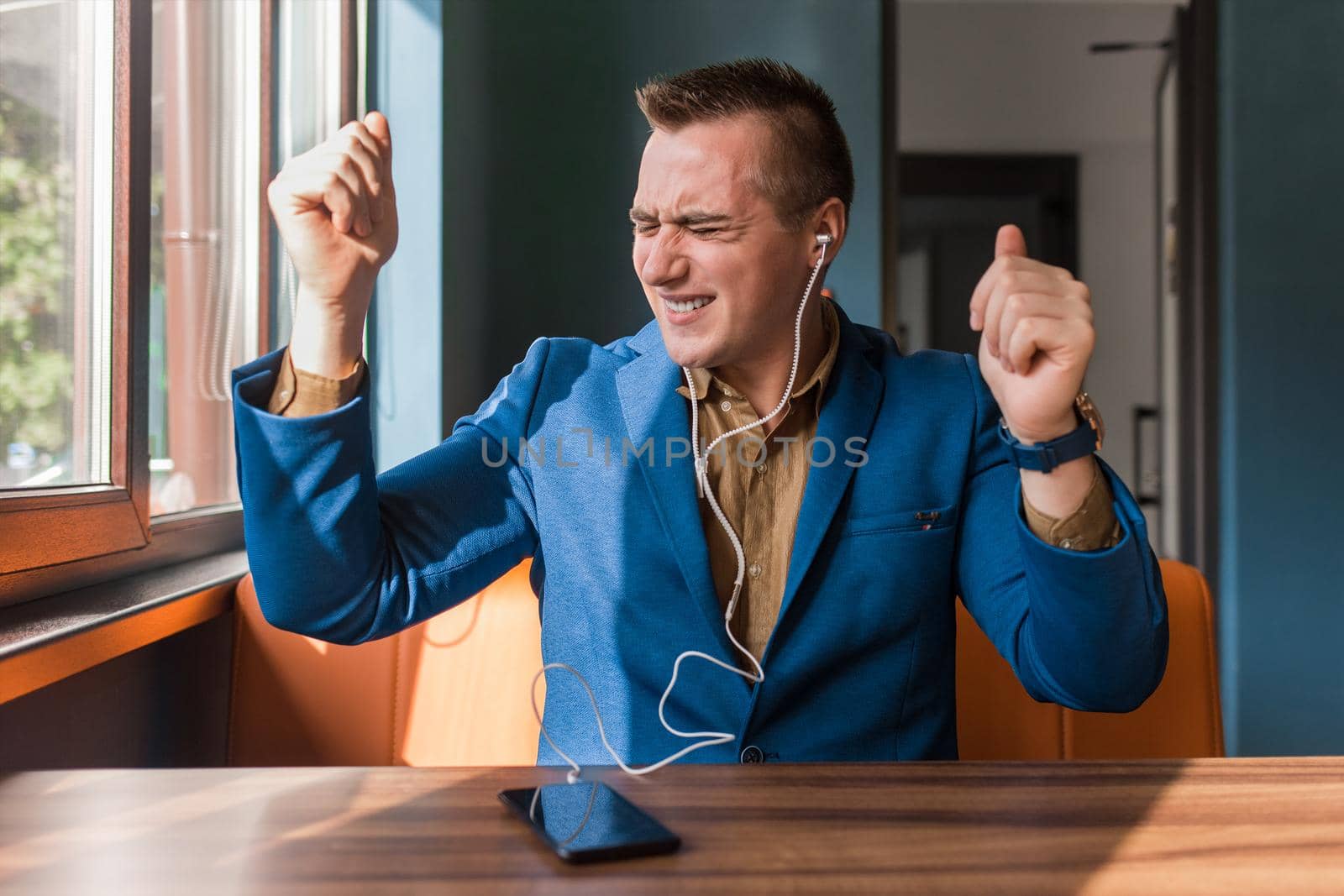 A handsome, carried away, funny dancing businessman of European appearance stylish portrait, holds a phone in his hand and listens to music in headphones sitting at a table in a cafe.