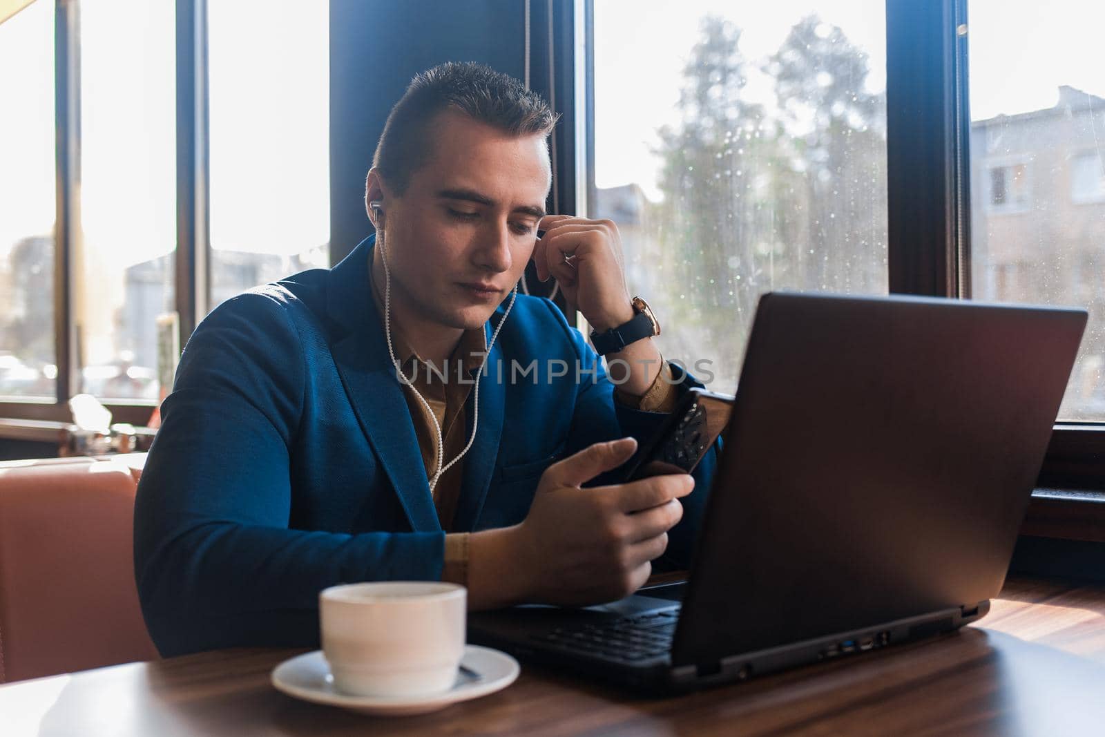 A business man stylish businessman in an attractive European-looking suit works in a laptop, listens to music with headphones and drinks coffee sitting at a table in a cafe by the window.