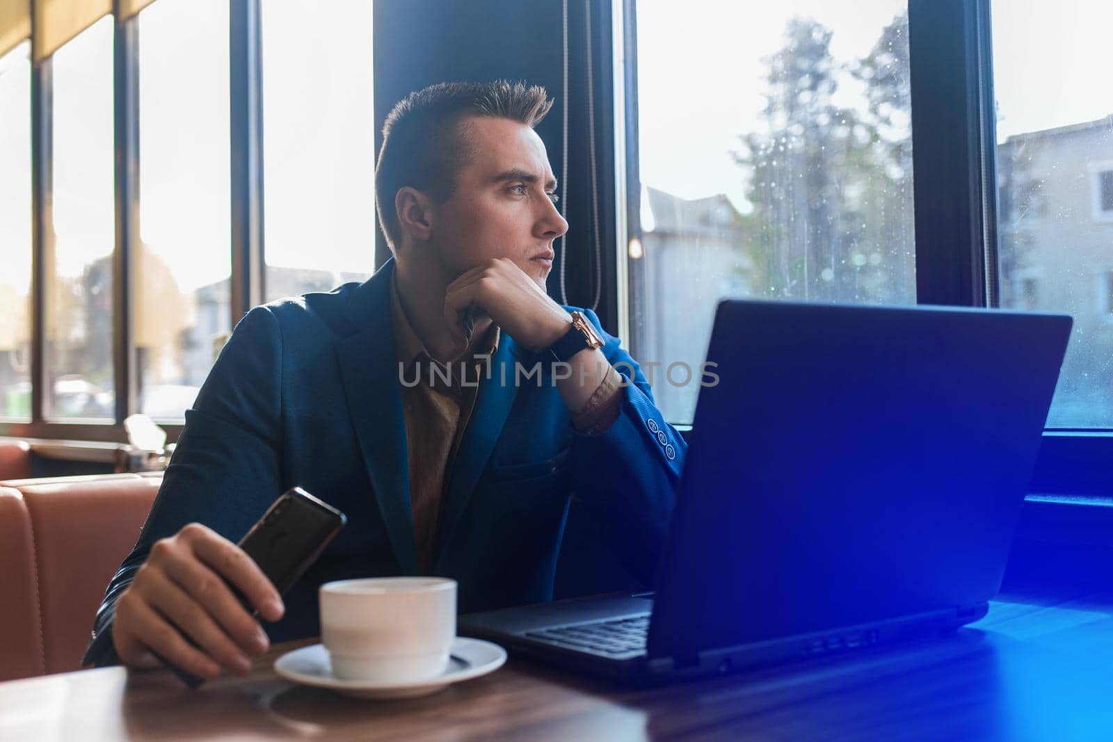 A business serious man pensive businessman a stylish of Caucasian appearance in a jacket, works in a laptop or computer, sitting at a table by the window in a cafe and holds mobile phone by AYDO8