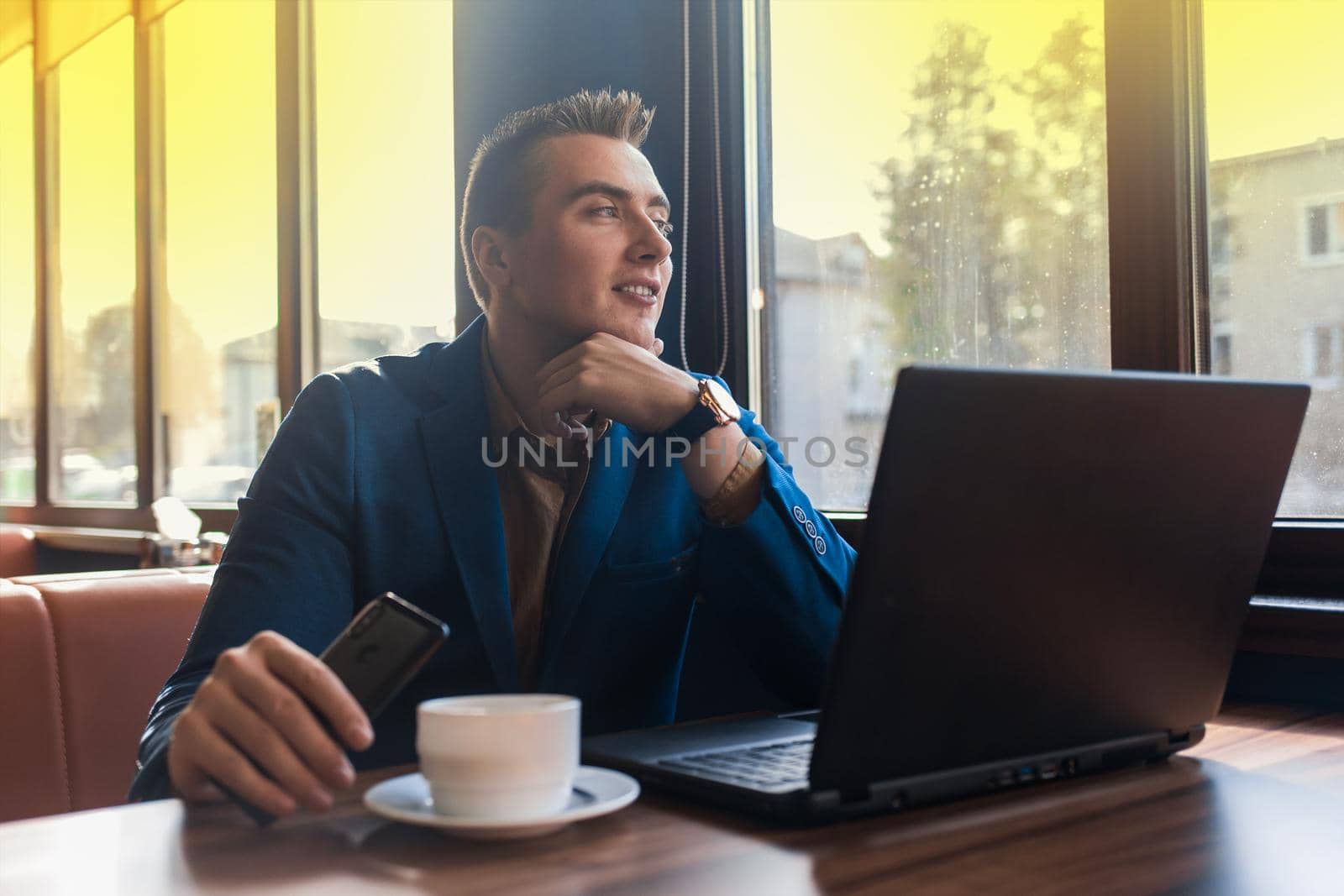 A business pleased man businessman a stylish of Caucasian appearance in a jacket, works in a laptop or computer, sitting at a table by the window in a cafe.