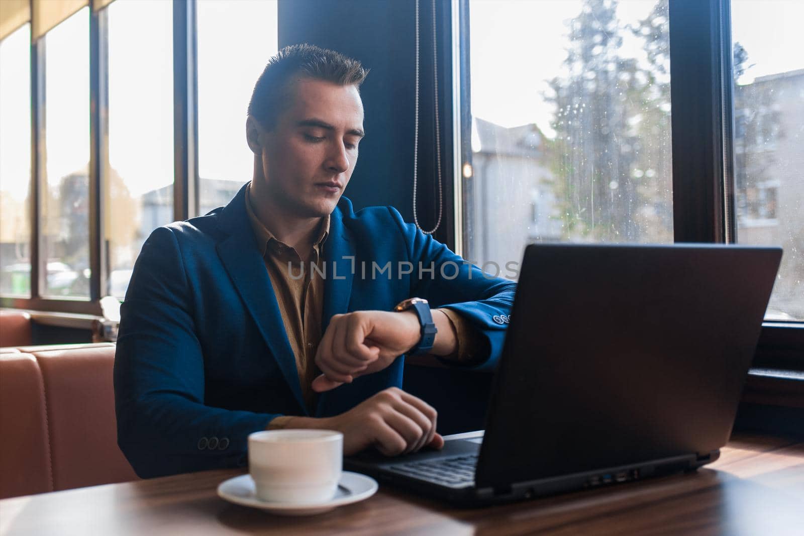 A business serious man businessman a stylish of Caucasian appearance in a jacket, works in a laptop or computer, sitting at a table by the window in a cafe and looks at the time on a wrist watch.