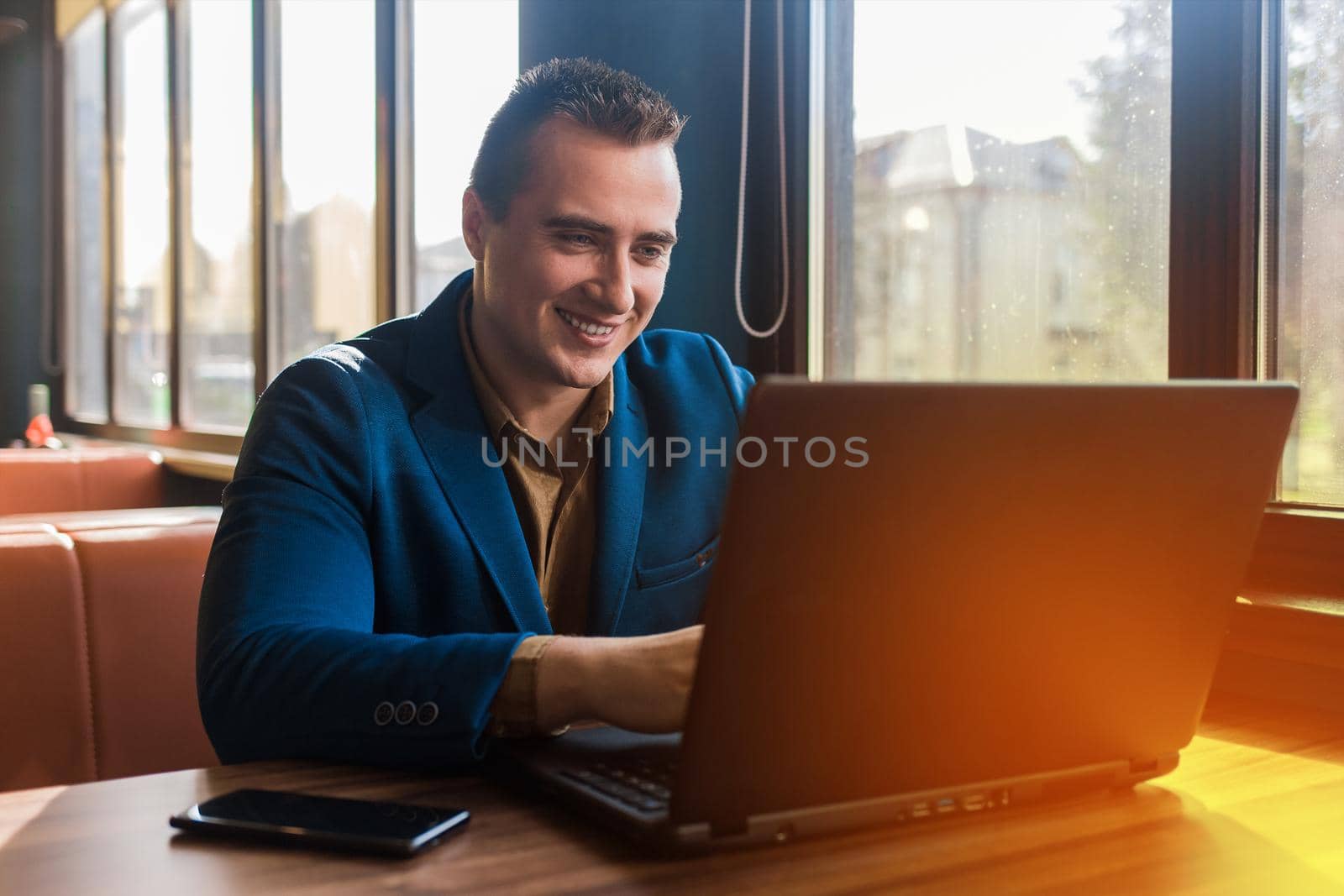 A business smiling man happy businessman a stylish portrait of Caucasian appearance in a jacket works in a laptop or computer, sitting at a table by the window in a cafe.